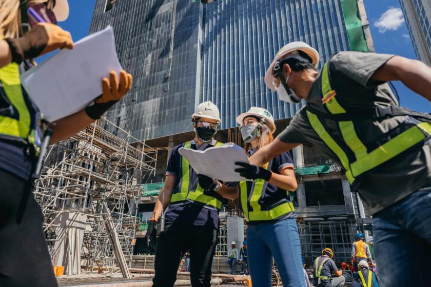 Construction workers in safety gear review blueprints at a building site, with scaffolding and a high-rise structure under construction in the background.