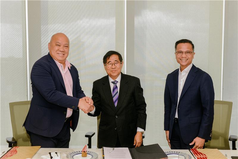 Three men in business attire stand together at a table. Two of them are shaking hands and smiling, while the third stands between them, also smiling. Documents are visible on the table, suggesting a formal agreement or partnership. The setting is a modern office with large windows in the background.