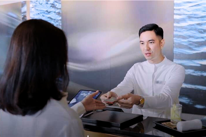 A hotel receptionist hands a key card to a guest across the front desk, both dressed in white, with a calm water-themed backdrop behind them.