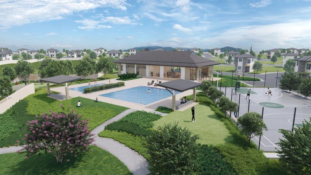 A landscaped community recreation area featuring a swimming pool, shaded lounging areas, and a sports court. The pool is surrounded by greenery, walking paths, and well-maintained lawns. A clubhouse with large windows overlooks the pool, while people are seen relaxing and enjoying the outdoor spaces. In the background, rows of modern houses are scattered throughout the neighborhood, with blue skies and mountains visible in the distance. The setting feels calm, inviting, and family-friendly.