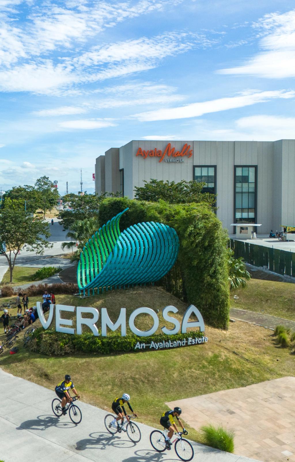 Cyclists ride past a large, modern sign that reads "Vermosa" in front of a landscaped area with lush greenery and a distinctive blue-and-green sculpture. In the background, a contemporary commercial building with the name "Ayala Malls" is visible under a bright, partly cloudy sky. The scene captures an active, urban lifestyle in a well-developed community space.