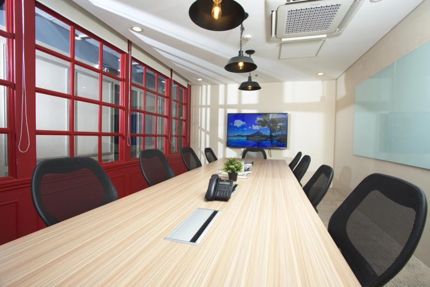 A modern conference room featuring a long wooden meeting table surrounded by black mesh office chairs. Overhead, three pendant lights hang from the ceiling. One wall is lined with bold red-framed windows, while the opposite wall has a mounted flat-screen TV displaying a scenic landscape. A glass whiteboard is mounted on another wall, and a phone and small plant sit on the center of the table.