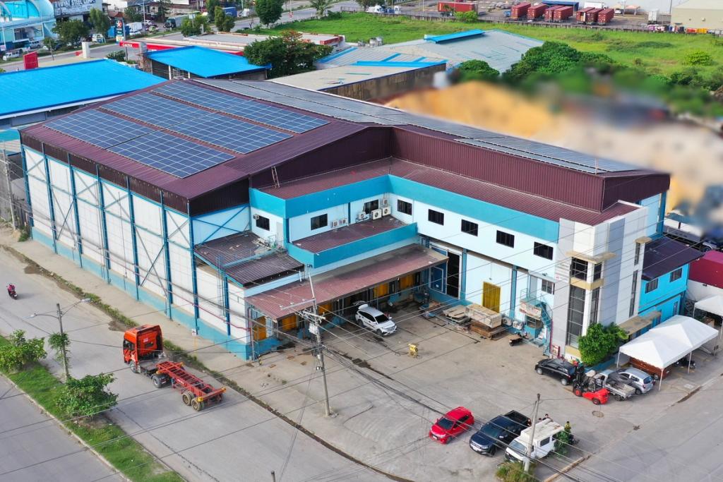 Aerial view of a modern industrial warehouse building with solar panels on the roof, loading dock area, parking space, and surrounding road infrastructure.