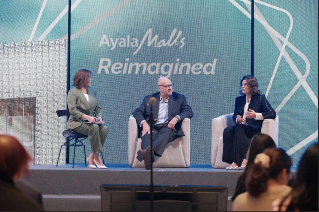 Three professionals are seated on stage in front of a large backdrop that reads "Ayala Malls Reimagined." They are engaged in a panel discussion, with one woman in a green suit, a man in a dark suit, and another woman in a black outfit. Audience members are visible in the foreground, listening attentively. The setting appears modern and formal, suitable for a corporate event or conference.