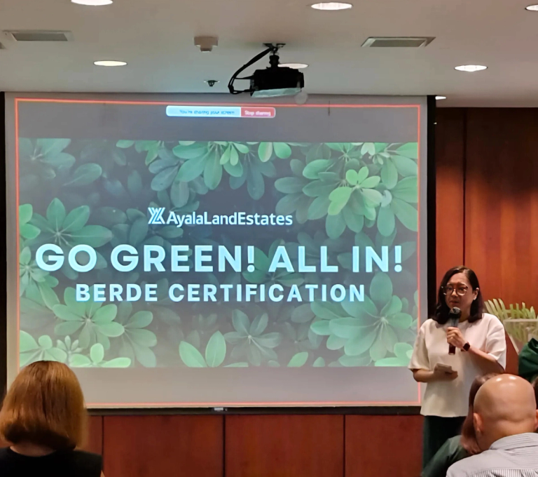 A woman stands and speaks in front of a group during a presentation. Behind her is a large screen displaying the Ayala Land Estates logo and the message: “GO GREEN! ALL IN! BERDE CERTIFICATION,” with a background of green leaves. The setting appears to be a conference or seminar room, with attendees seated and listening.
