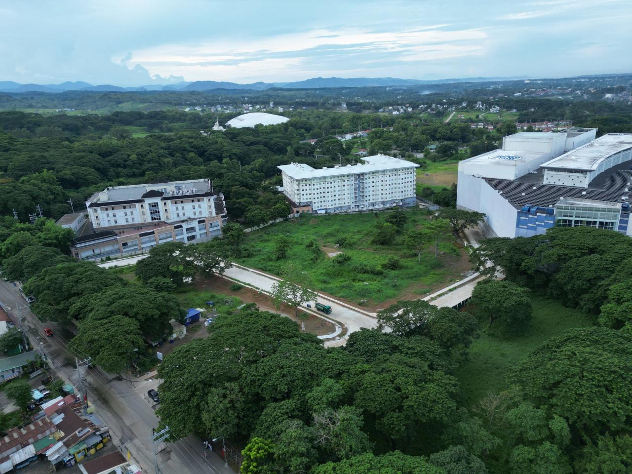 Aerial view of a semi-urban area featuring large institutional buildings surrounded by dense greenery and trees. In the center, there’s an open grassy plot with newly constructed paved roads. Residential neighborhoods and forested hills extend into the distance under a cloudy sky, with mountains visible on the horizon.