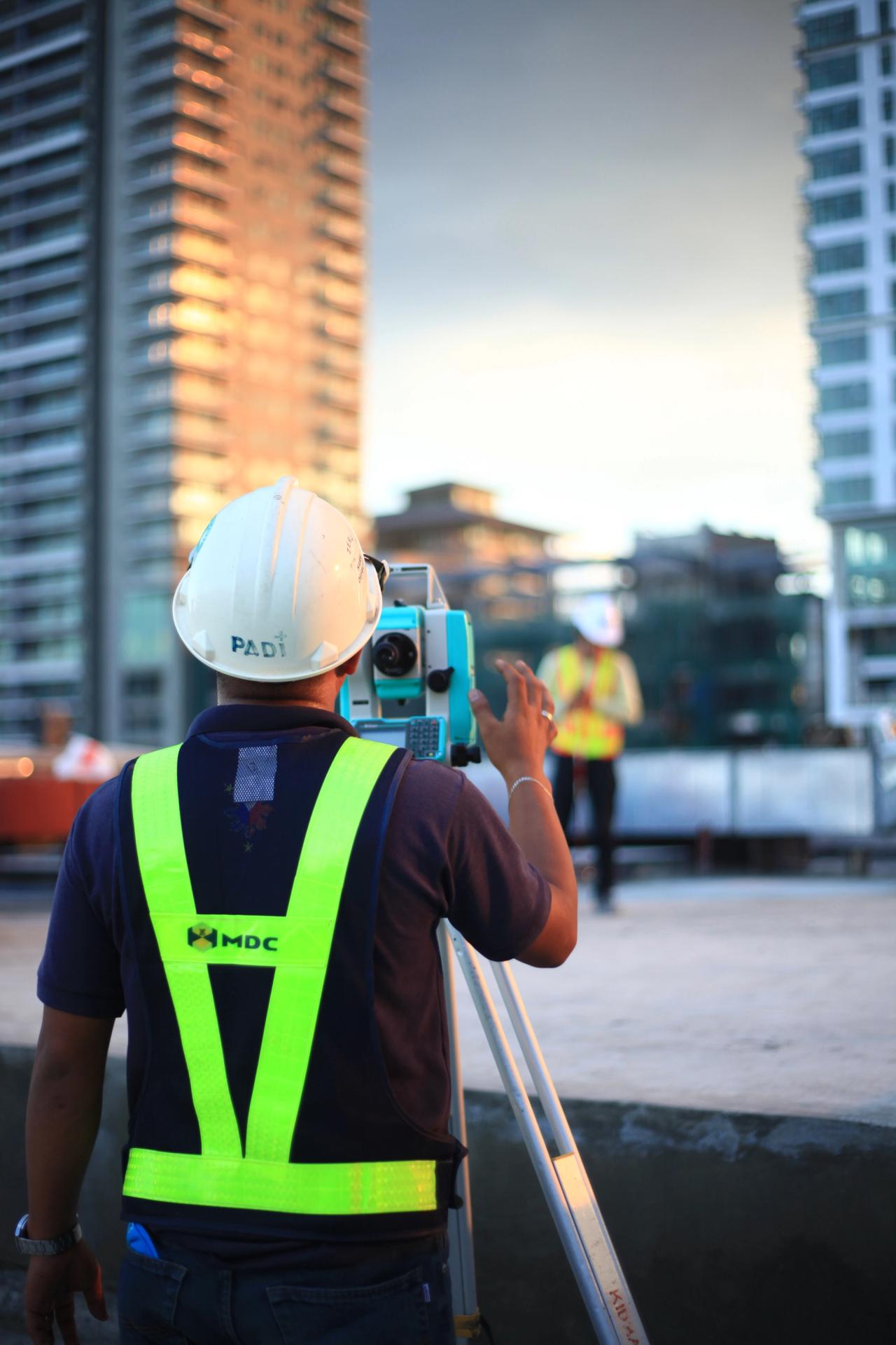 A construction worker in a hard hat and high-visibility vest operates a surveying instrument on a building site, with high-rise buildings in the background during sunset.