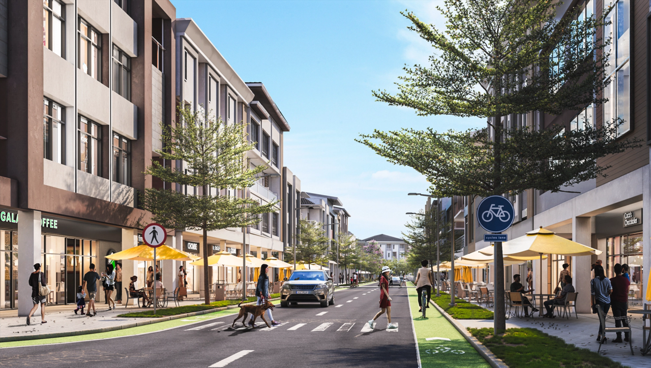 Street-level view of a walkable, mixed-use district with mid-rise buildings, outdoor cafés shaded by yellow umbrellas, bike lanes, and people enjoying a vibrant, pedestrian-friendly environment under a clear blue sky.