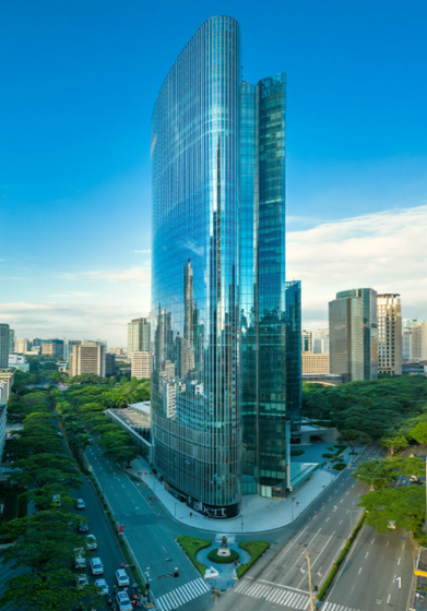 A tall, curved glass skyscraper rises above a busy city intersection, reflecting the surrounding buildings and blue sky. Trees line the streets below.