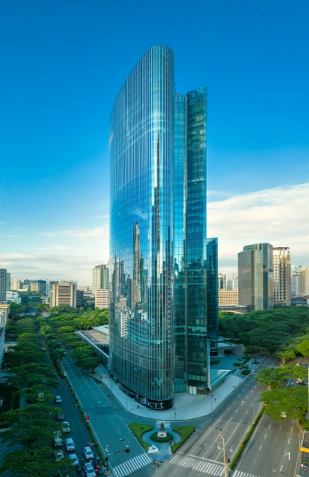 A tall, curved glass skyscraper rises above a busy city intersection, reflecting the surrounding buildings and blue sky. Trees line the streets below