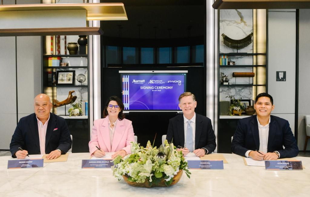 Four business professionals, three men and one women, are seated at a marble table, each signing documents. They are dressed in formal attire and smiling at the camera. In the background, a large screen displays the logos of Marriott, AyalaLand Hospitality, and Moxy, along with the words "Signing Ceremony." The setting appears modern and professional, with shelves displaying books, decor, and plants. A floral arrangement sits in the center of the table.
