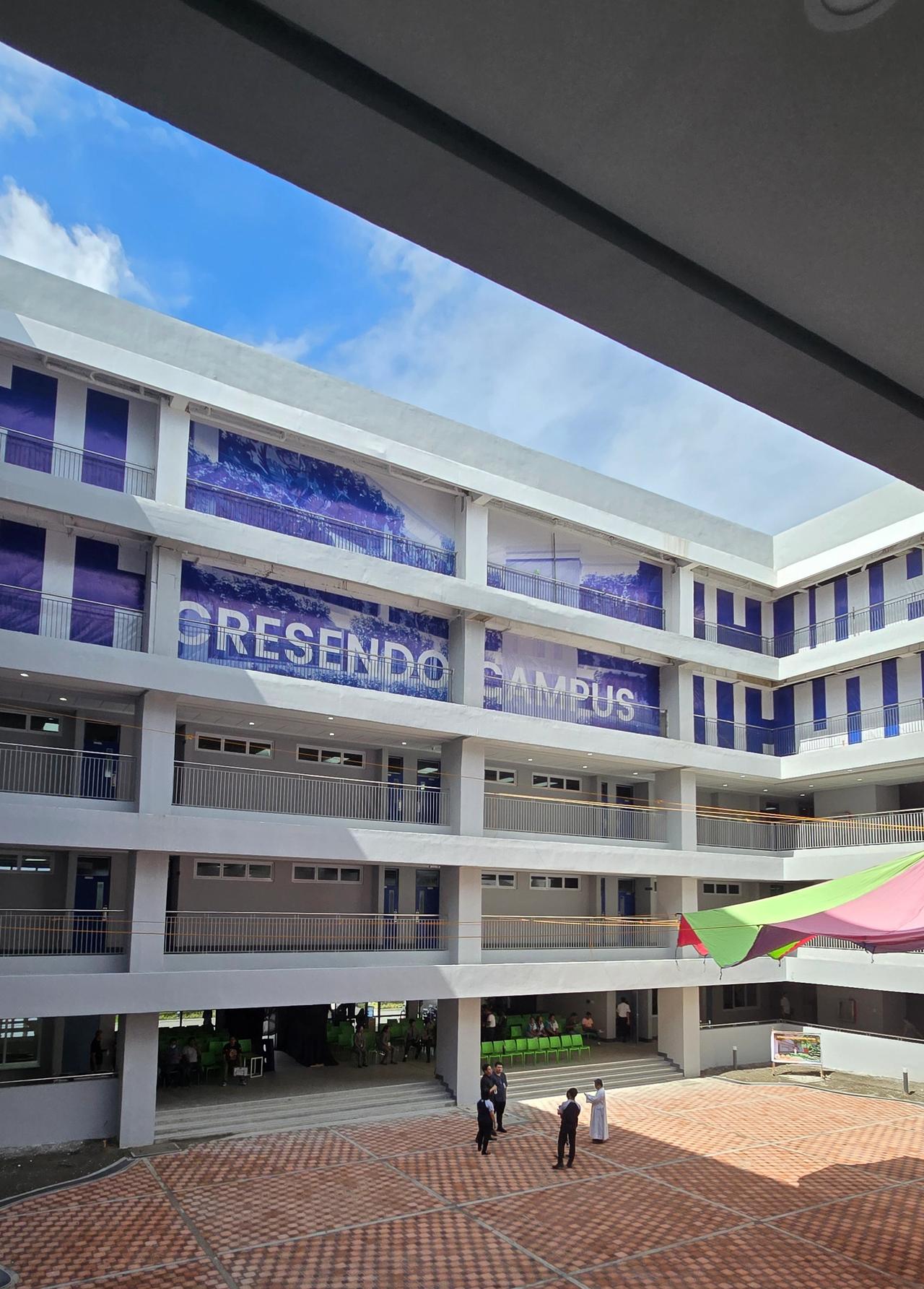 Modern multi-story educational building labeled 'Cresendo Campus' with open-air hallways and a central courtyard. A few people stand on the red-brick ground level under a partly cloudy blue sky, viewed from beneath a shaded overhang.