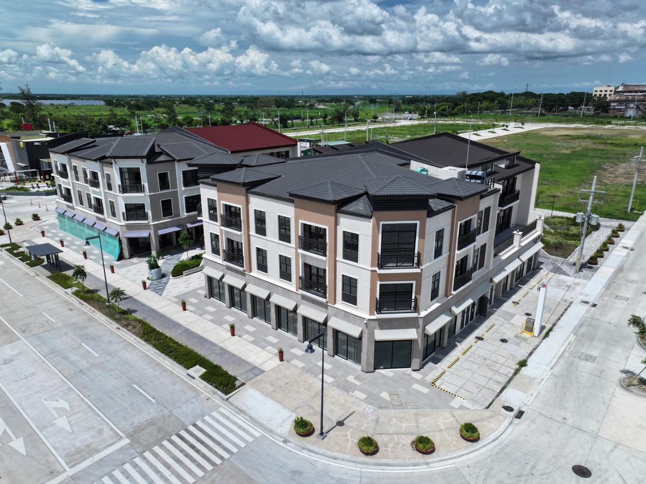Aerial view of a newly constructed, modern three-story commercial building complex with empty storefronts on the ground level. The buildings feature dark roofs, large windows, and clean architectural lines. Surrounding the complex are wide roads, sidewalks, and open plots of undeveloped land under a partly cloudy sky.