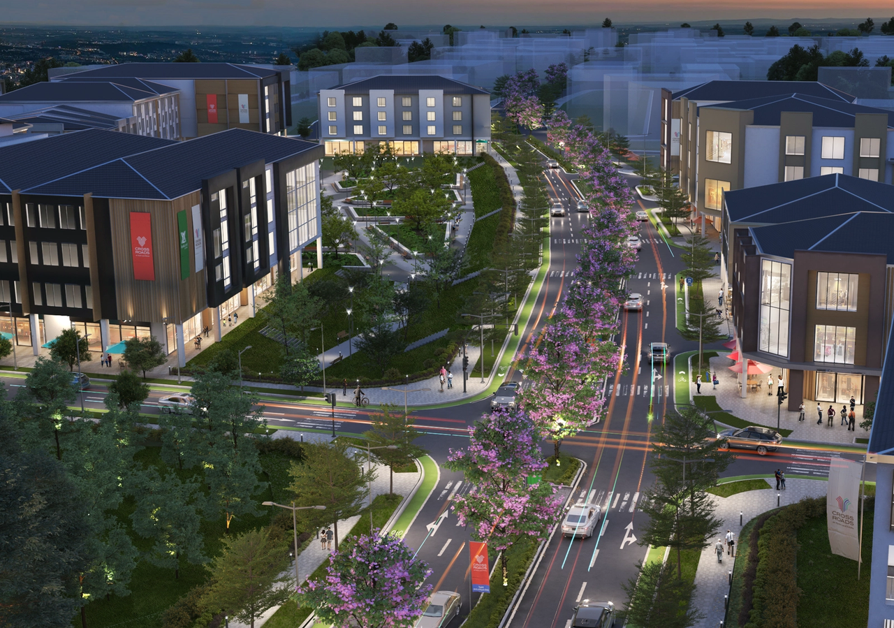 Evening aerial view of a well-lit, tree-lined boulevard in a modern mixed-use community, featuring mid-rise residential and commercial buildings, landscaped walkways, vibrant retail spaces, and blooming street trees under a twilight sky.