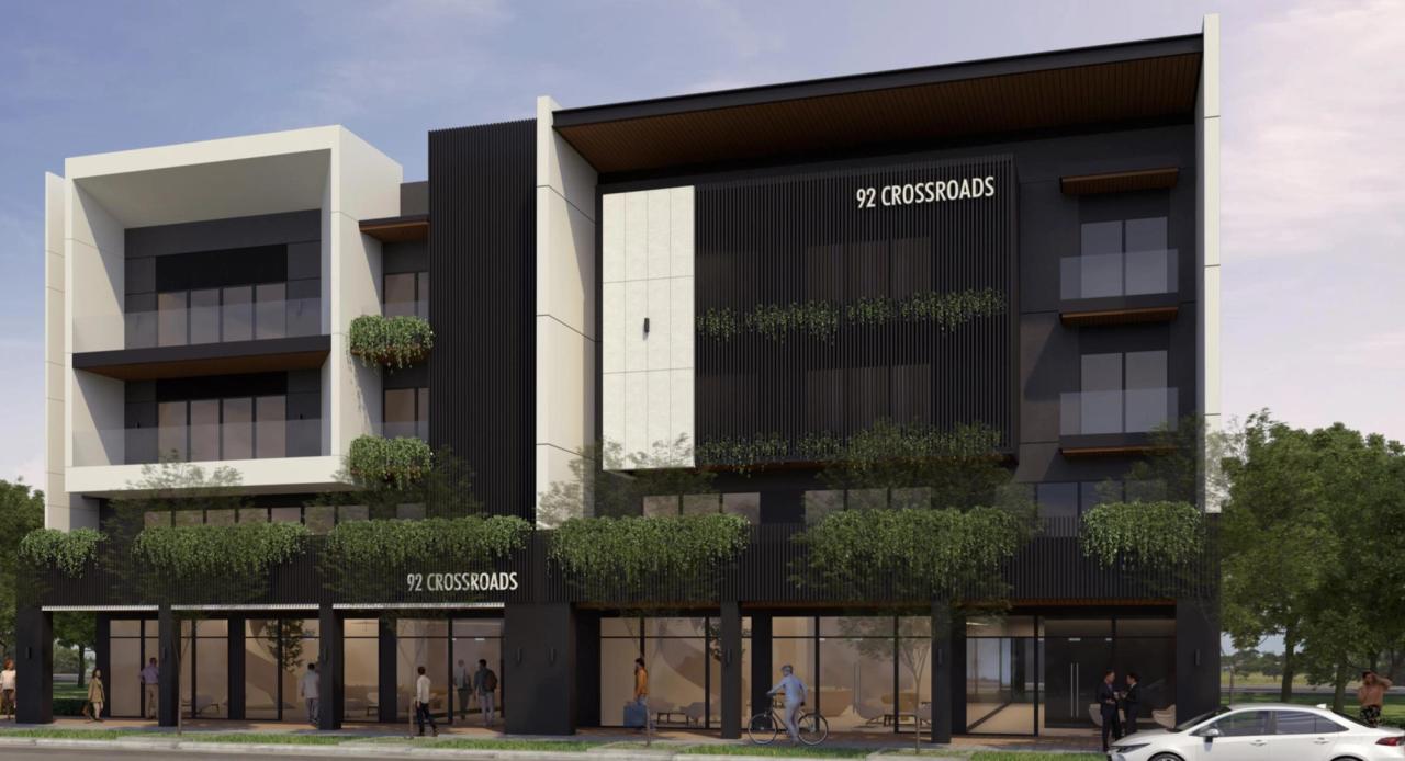 Architectural rendering of a modern mid-rise commercial building named “92 Crossroads,” featuring clean geometric lines, black and white façade elements, glass balconies, and greenery integrated into the design. The ground floor includes glass storefronts with visible activity, set against a backdrop of trees and open sky.