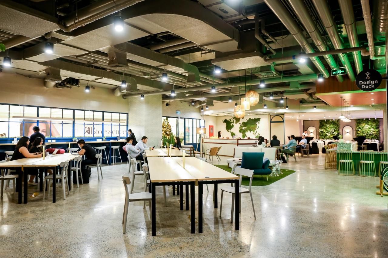 A spacious, modern coworking area with open seating, long tables, and people working or relaxing. The space features bright lighting, and exposed ceilings for an industrial look.