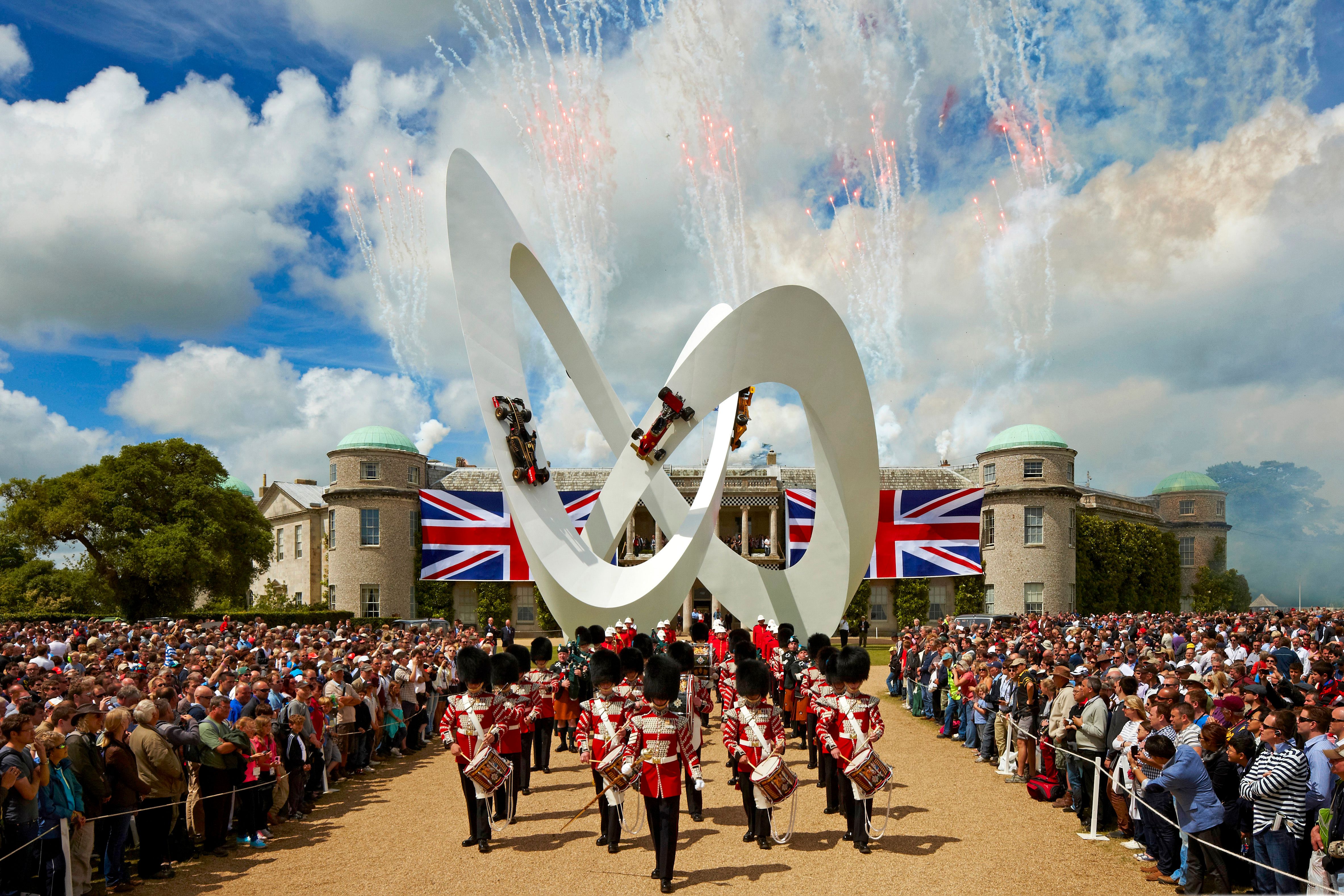 A ceremonial band marches past a white sculpture with F1 cars, a large crowd, a grand house with Union Jack flags, and fireworks.