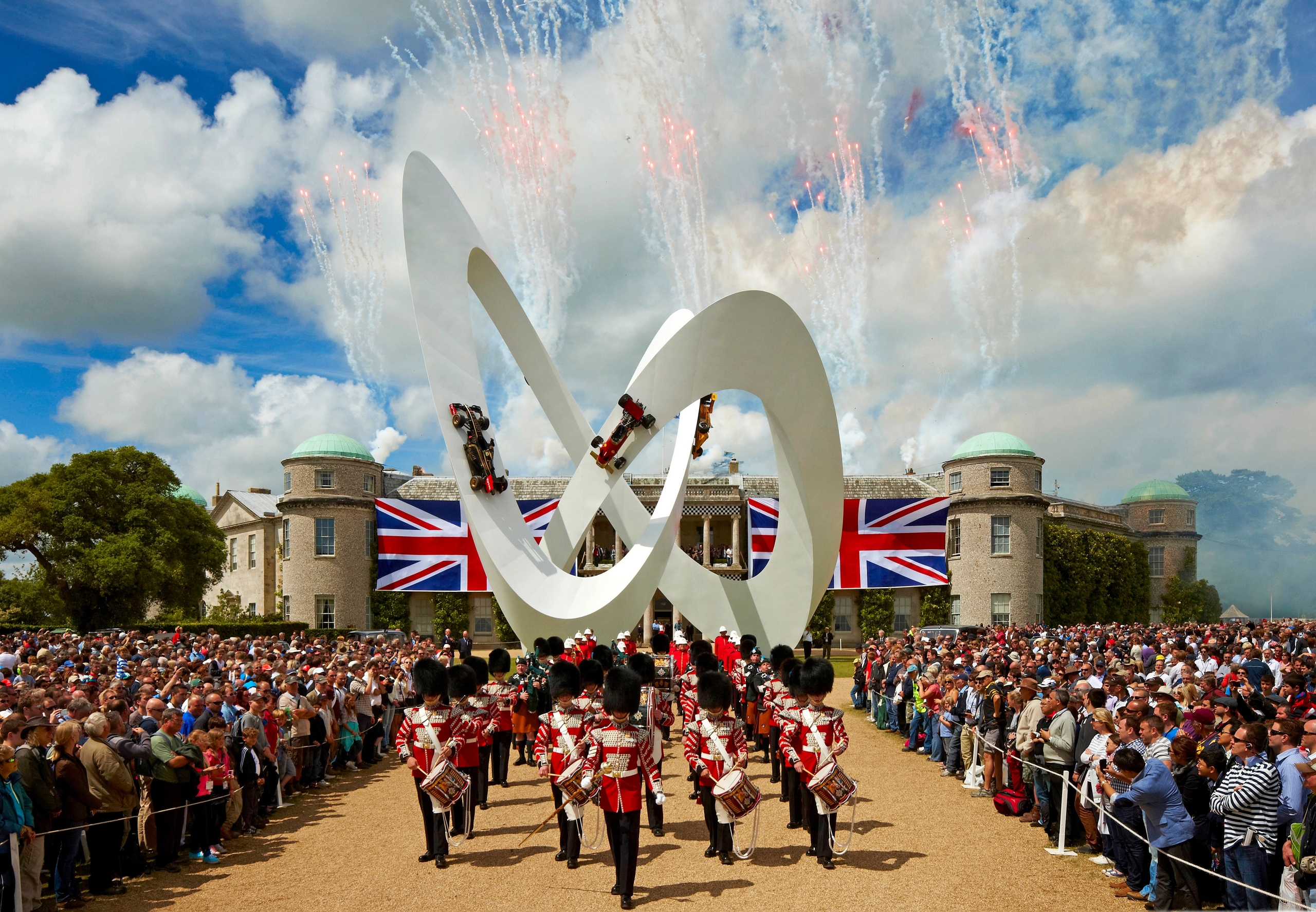 A ceremonial band marches past a white sculpture with F1 cars, a large crowd, a grand house with Union Jack flags, and fireworks.