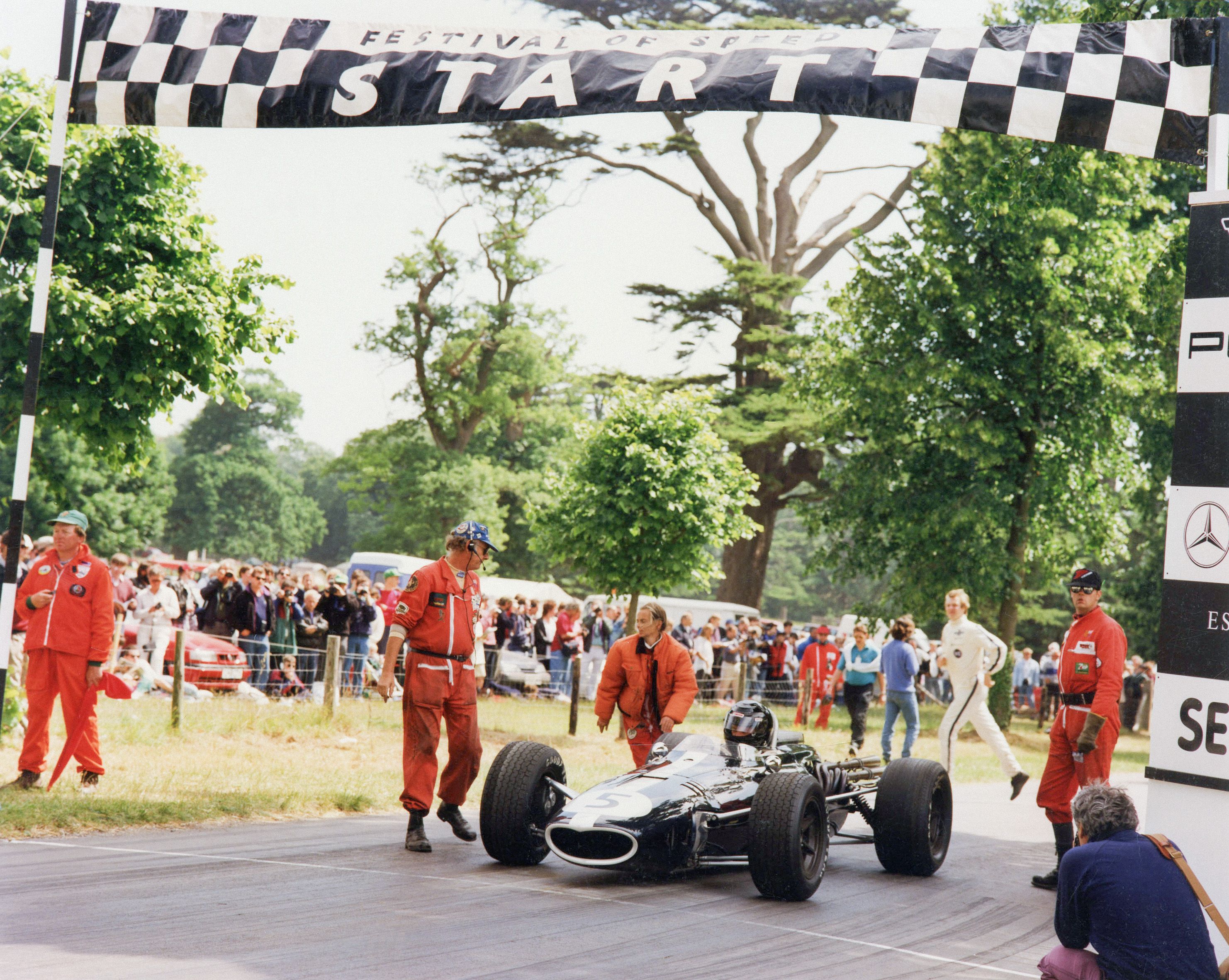 A classic black F1 car, number 5, at a 'START' line with crew and spectators at an outdoor event.