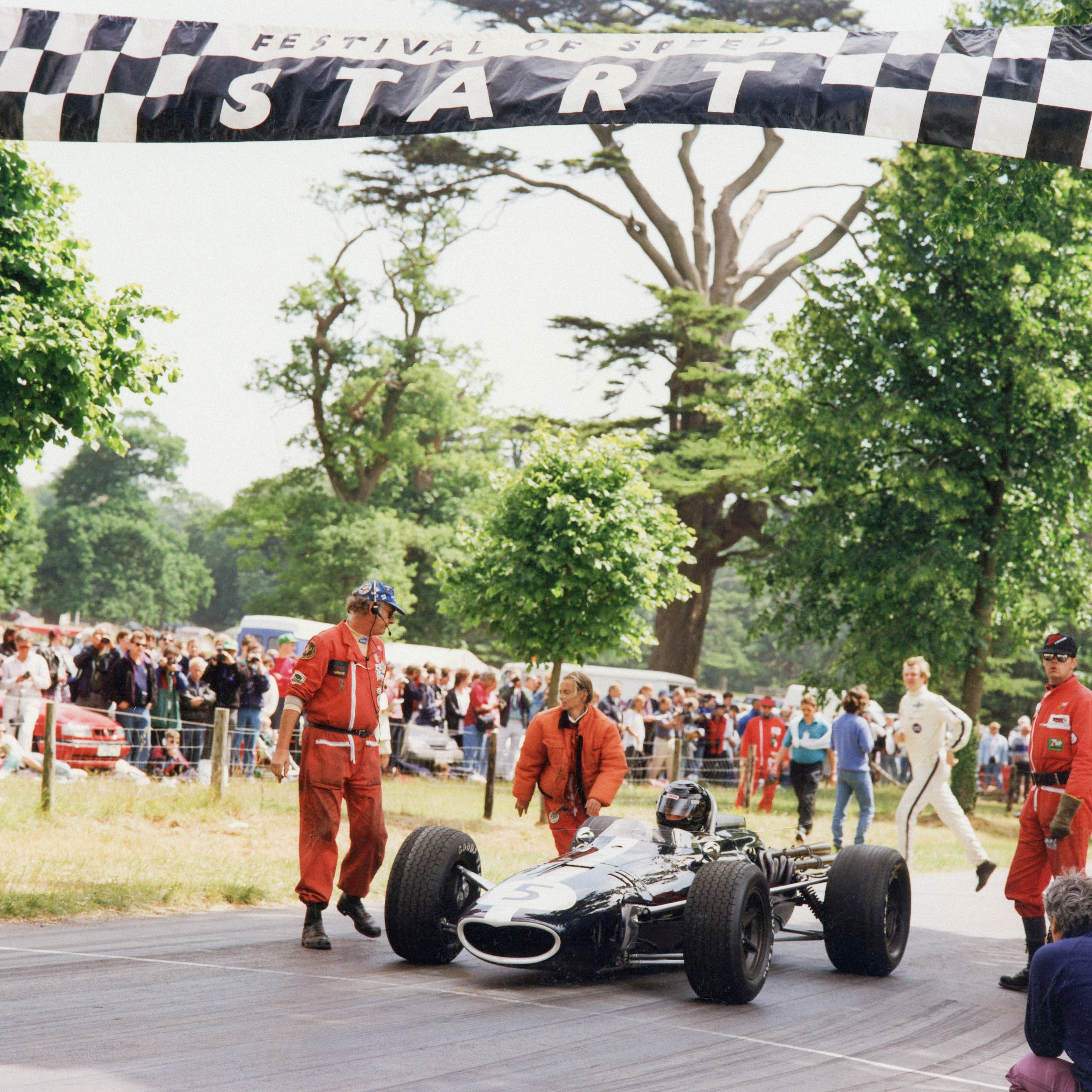 A classic black F1 car, number 5, at a 'START' line with crew and spectators at an outdoor event.