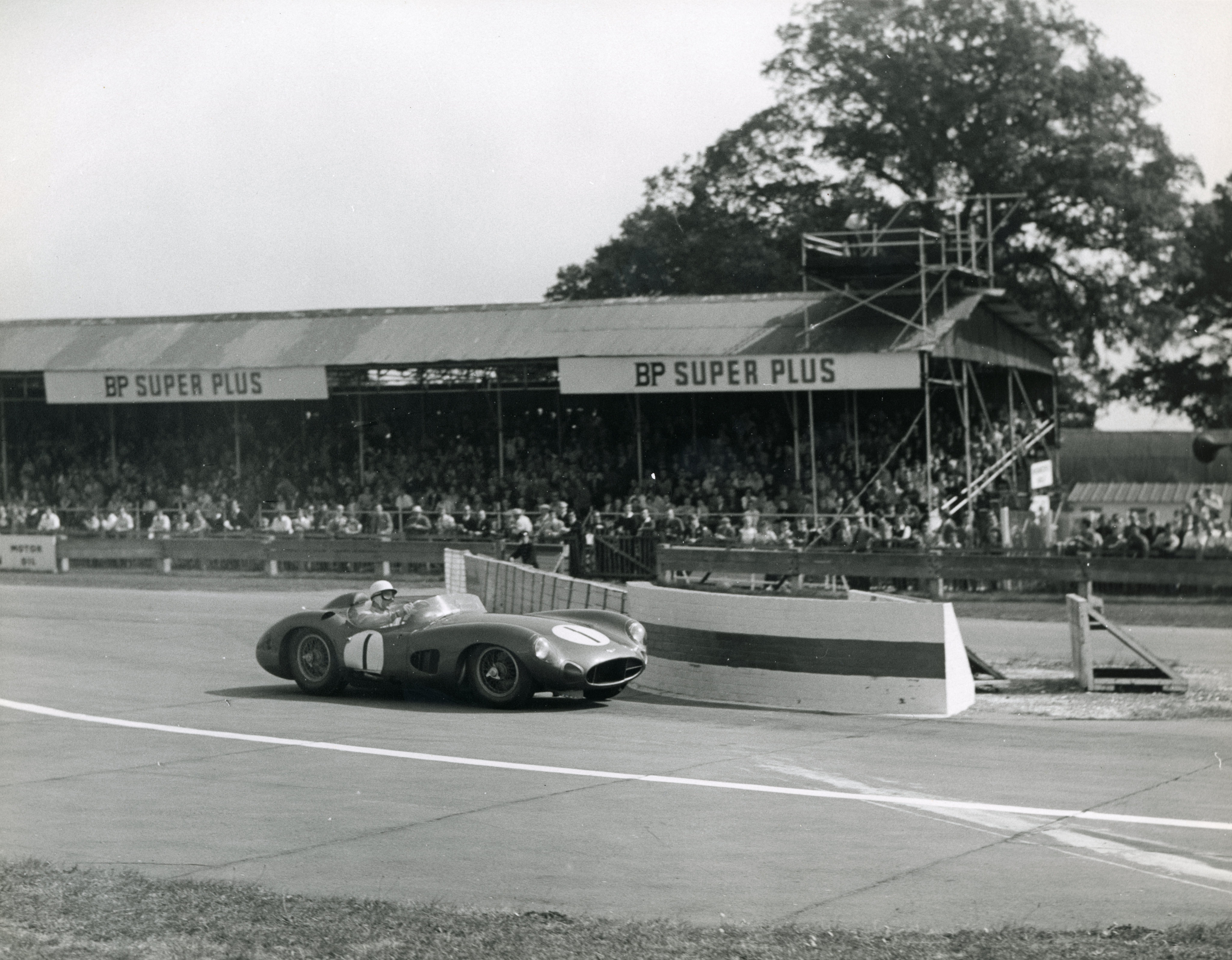 Black and white photo of a vintage race car with number 1 rounding a turn on a track, with a grandstand full of spectators behind it.