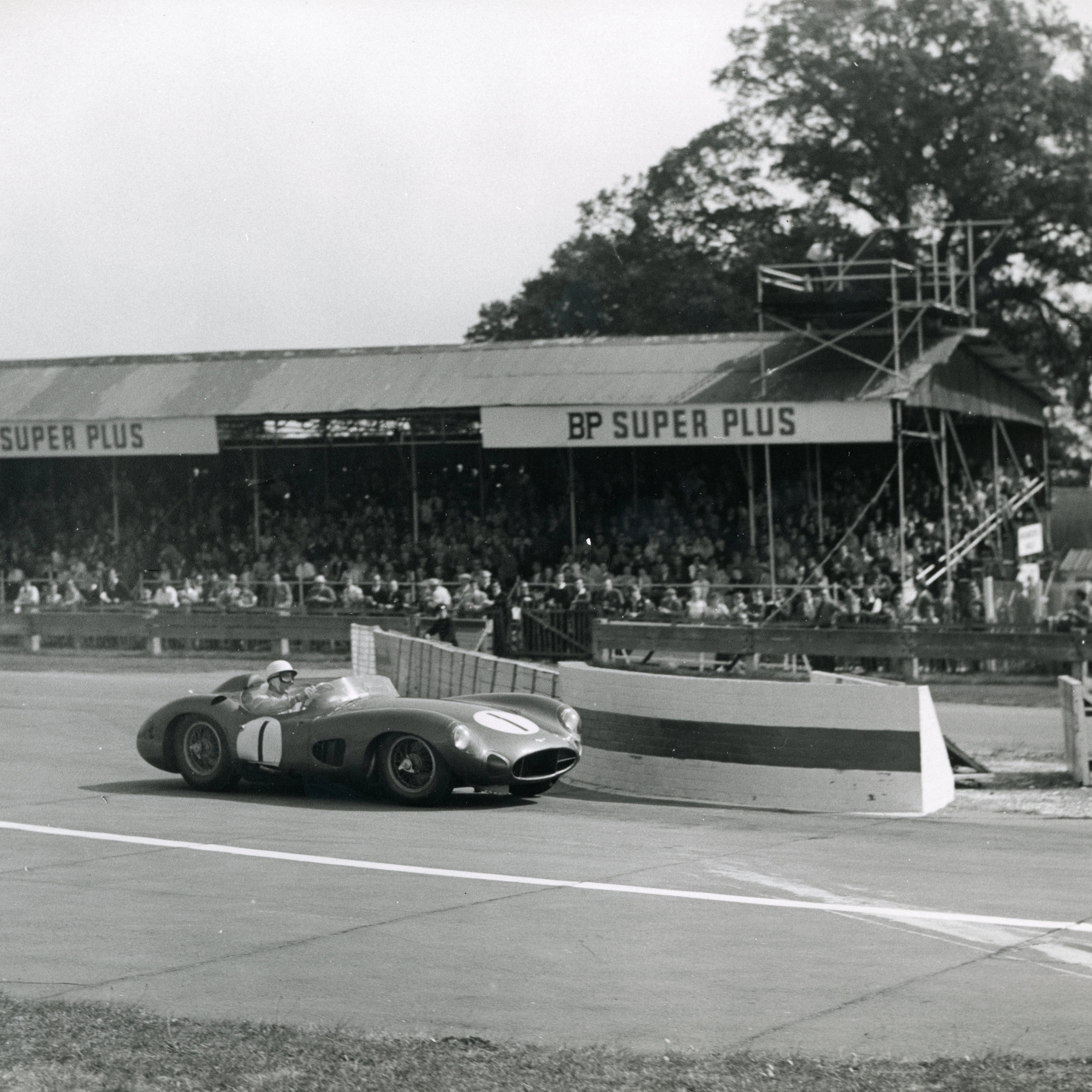 Black and white photo of a vintage race car with number 1 rounding a turn on a track, with a grandstand full of spectators behind it.