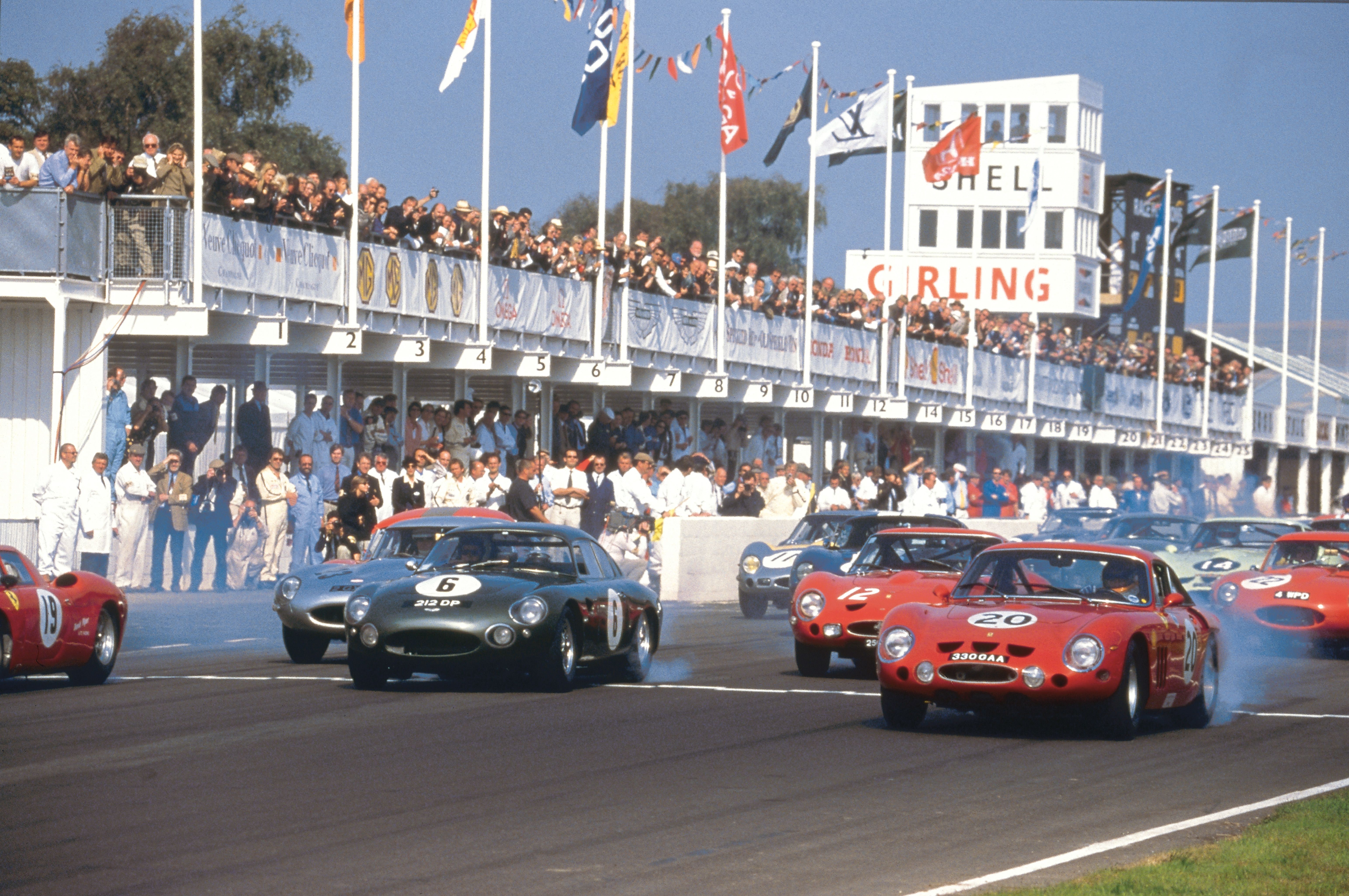 Vintage sports cars kicking up smoke at the start of a race, with spectators and a control tower in the background.
