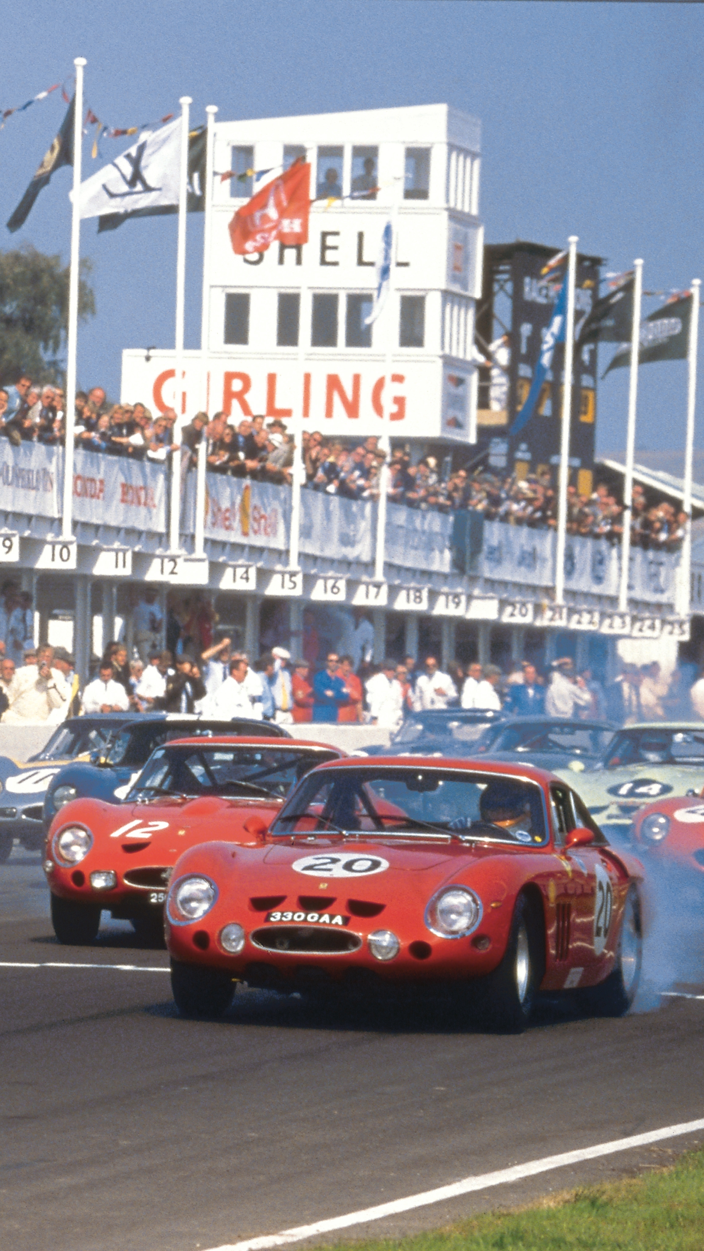 Vintage sports cars kicking up smoke at the start of a race, with spectators and a control tower in the background.