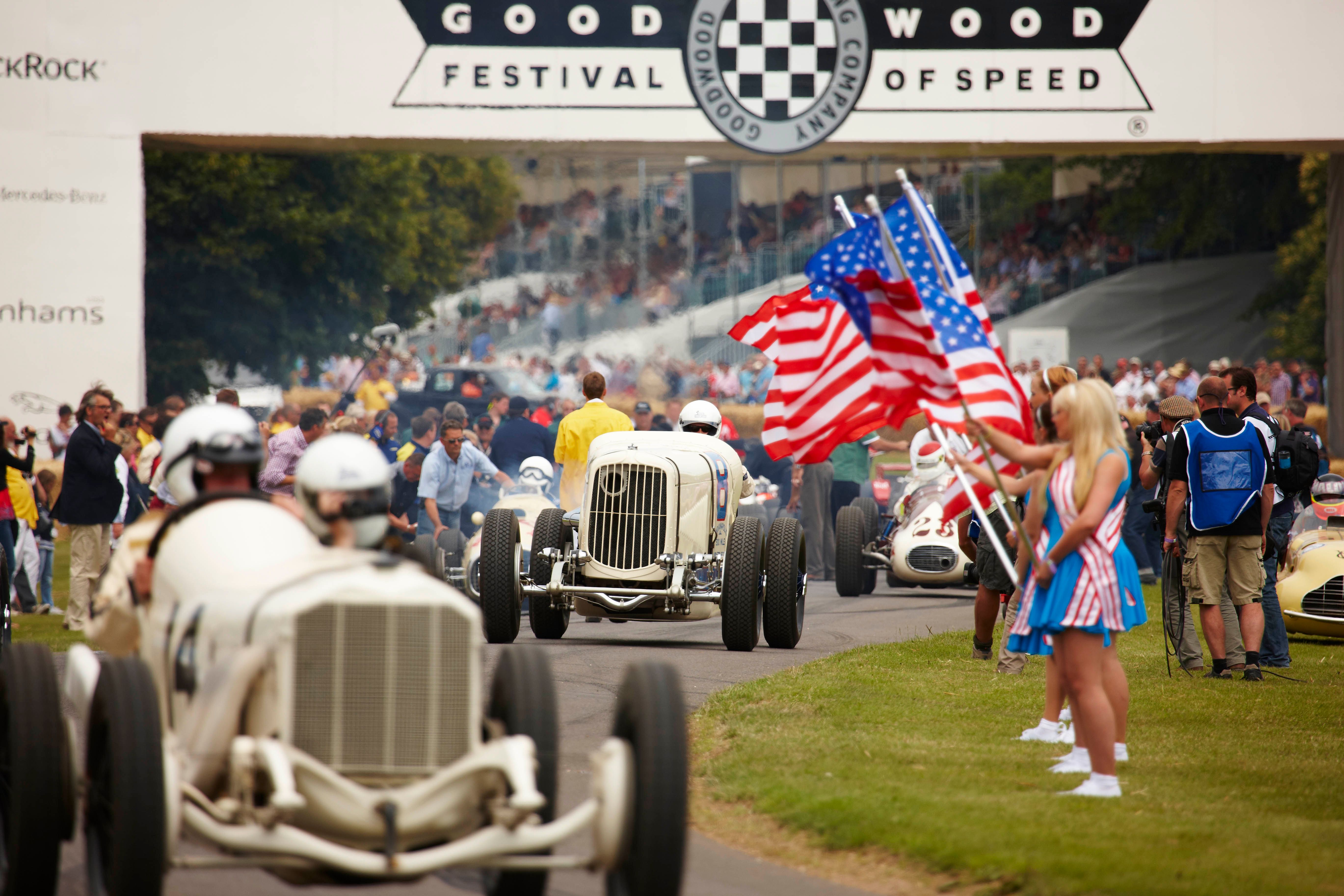 A marching band carrying American flags parades under a "Goodwood Festival of Speed" banner, with classic cars and spectators in the background.