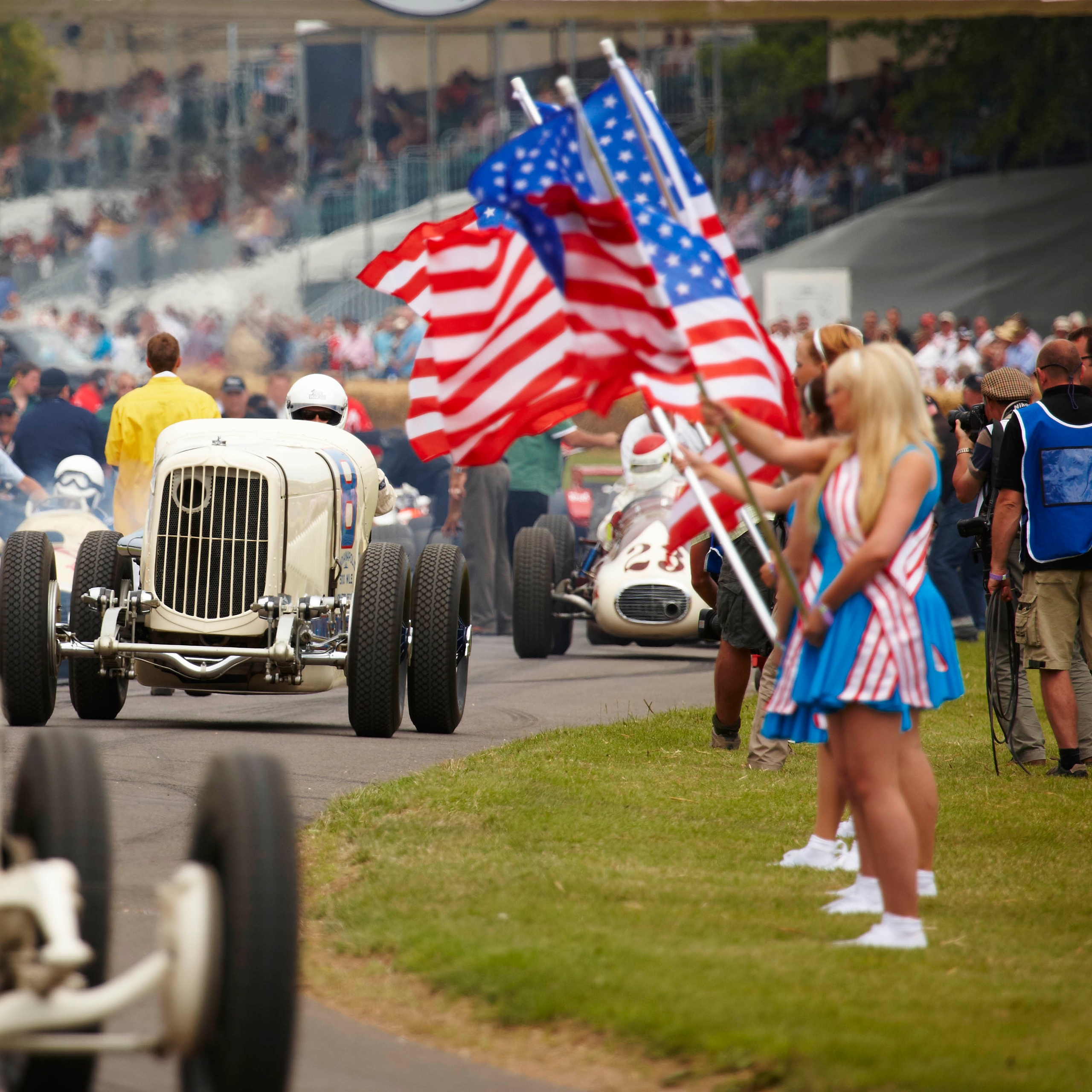 A marching band carrying American flags parades under a "Goodwood Festival of Speed" banner, with classic cars and spectators in the background.