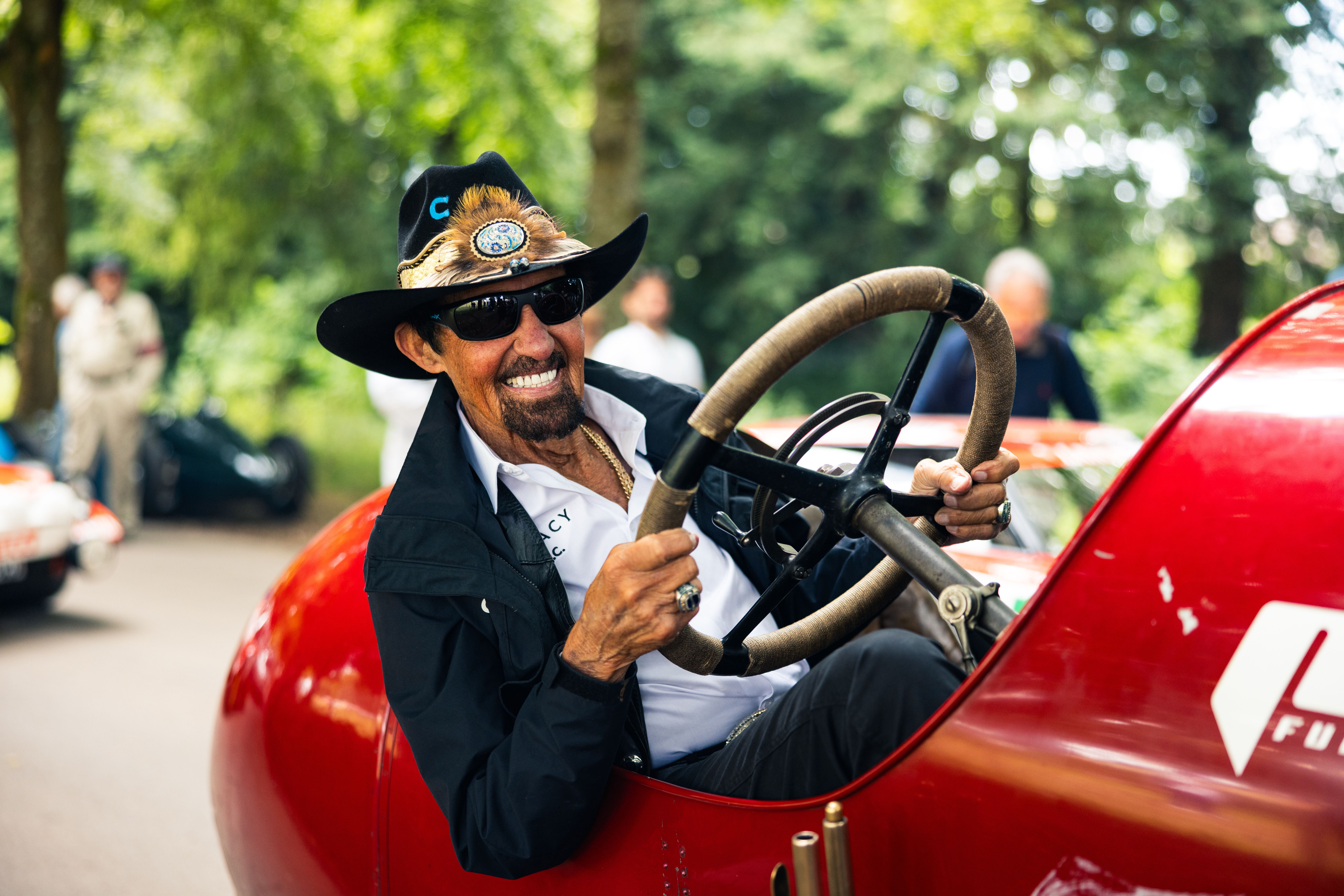 Richard Petty in a cowboy hat and sunglasses, behind the wheel of a red vintage race car.