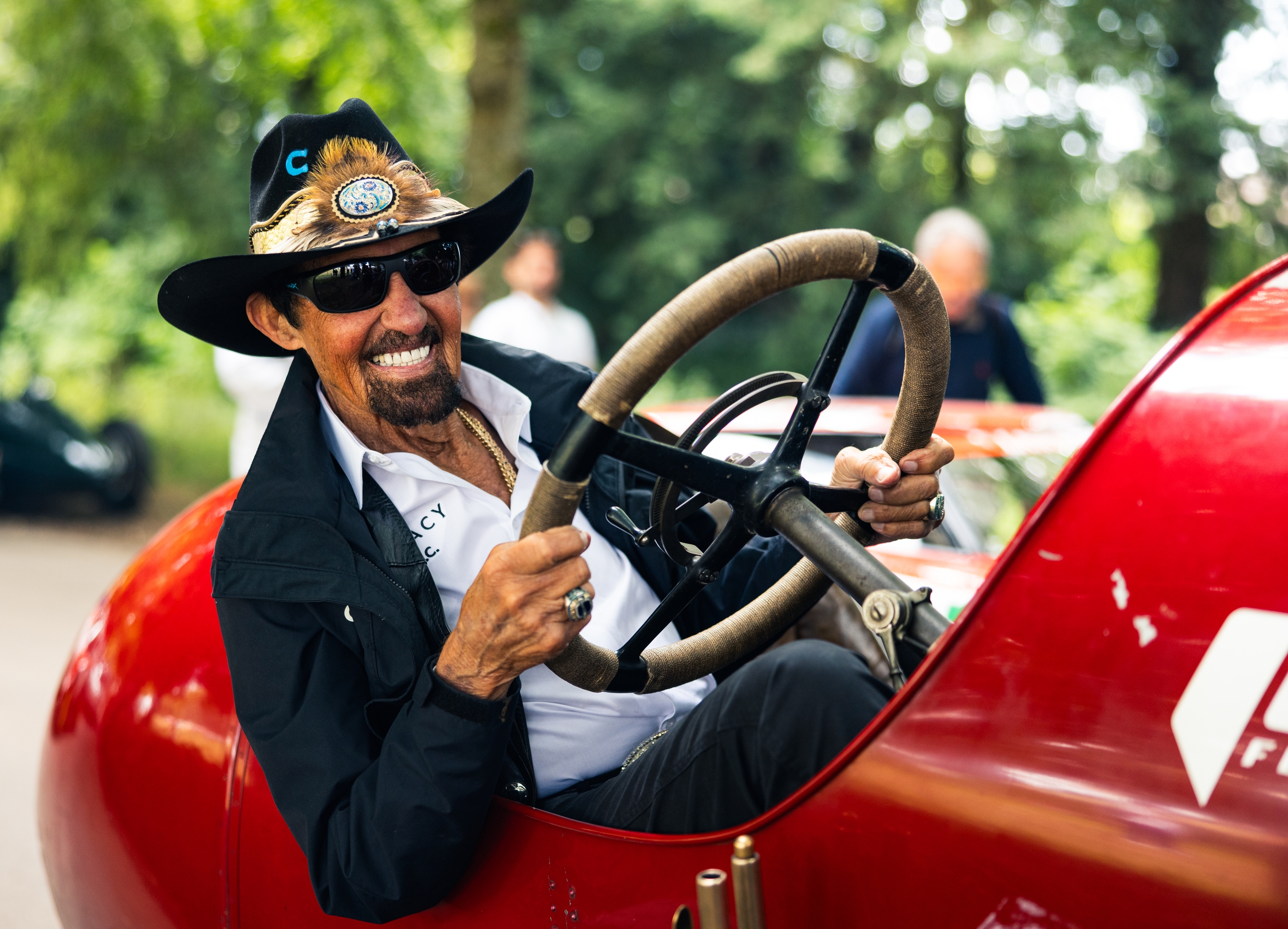 Richard Petty in a cowboy hat and sunglasses, behind the wheel of a red vintage race car.
