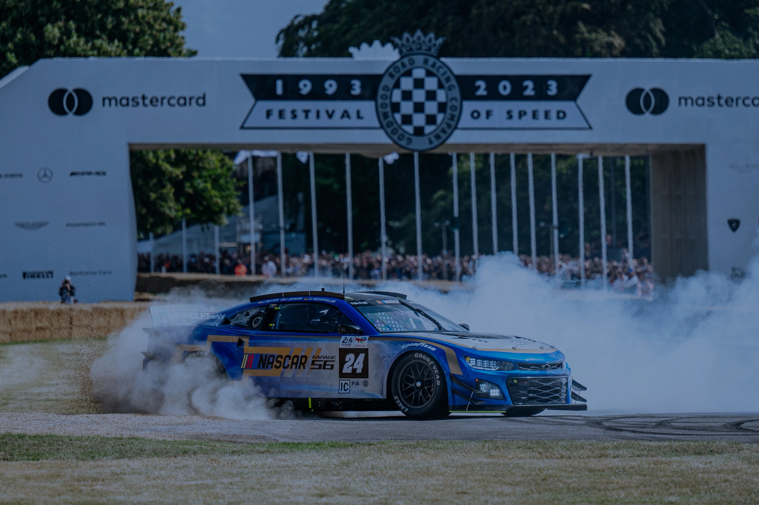A red race car with sponsors drives past a crowd of spectators at an outdoor event.