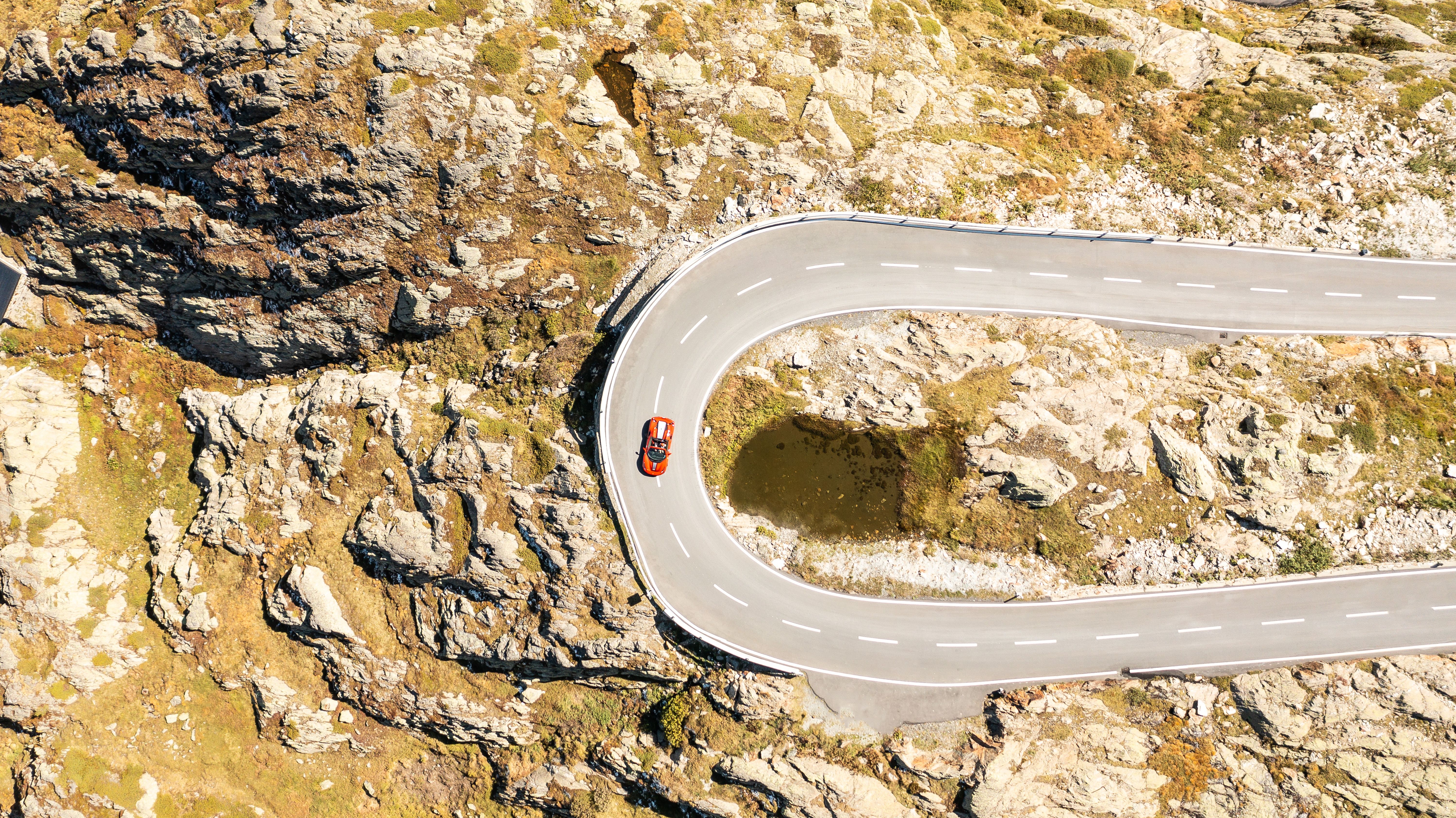 A light-colored vintage Porsche drives quickly on a winding road through green hills under a partly cloudy sky.