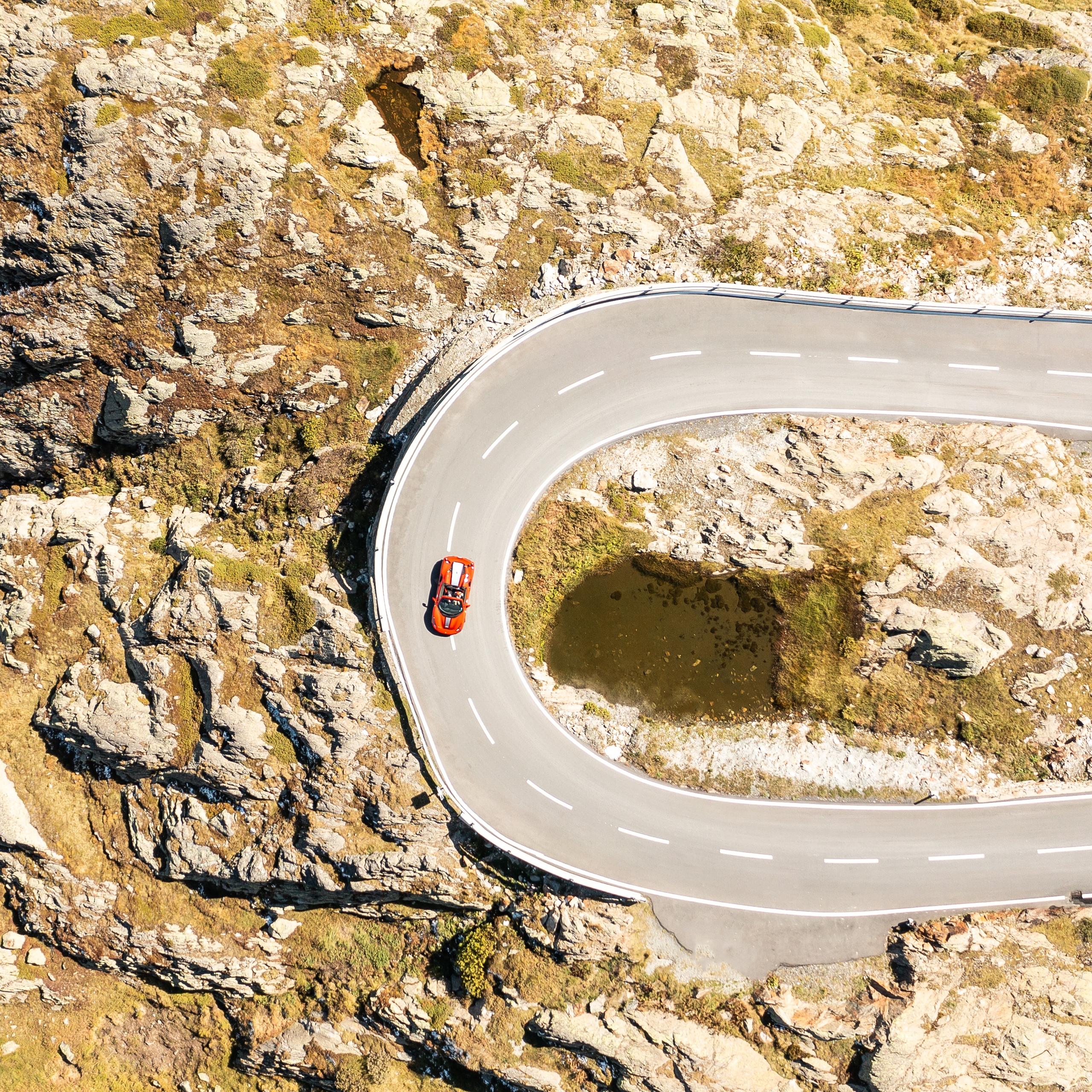 A light-colored vintage Porsche drives quickly on a winding road through green hills under a partly cloudy sky.