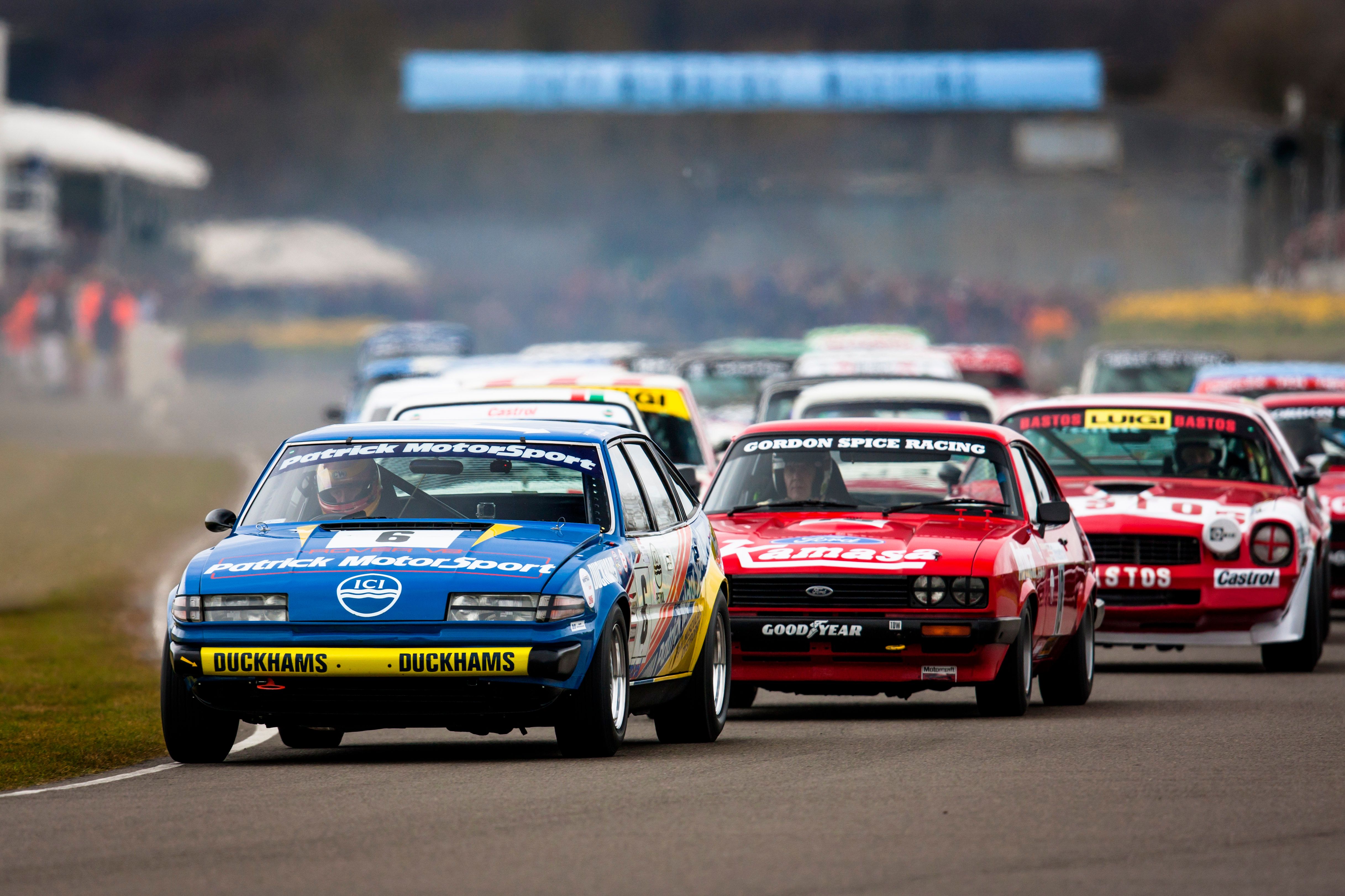 Vintage race cars on a track, with spectators and hills in the background at the 77th Members' Meeting.