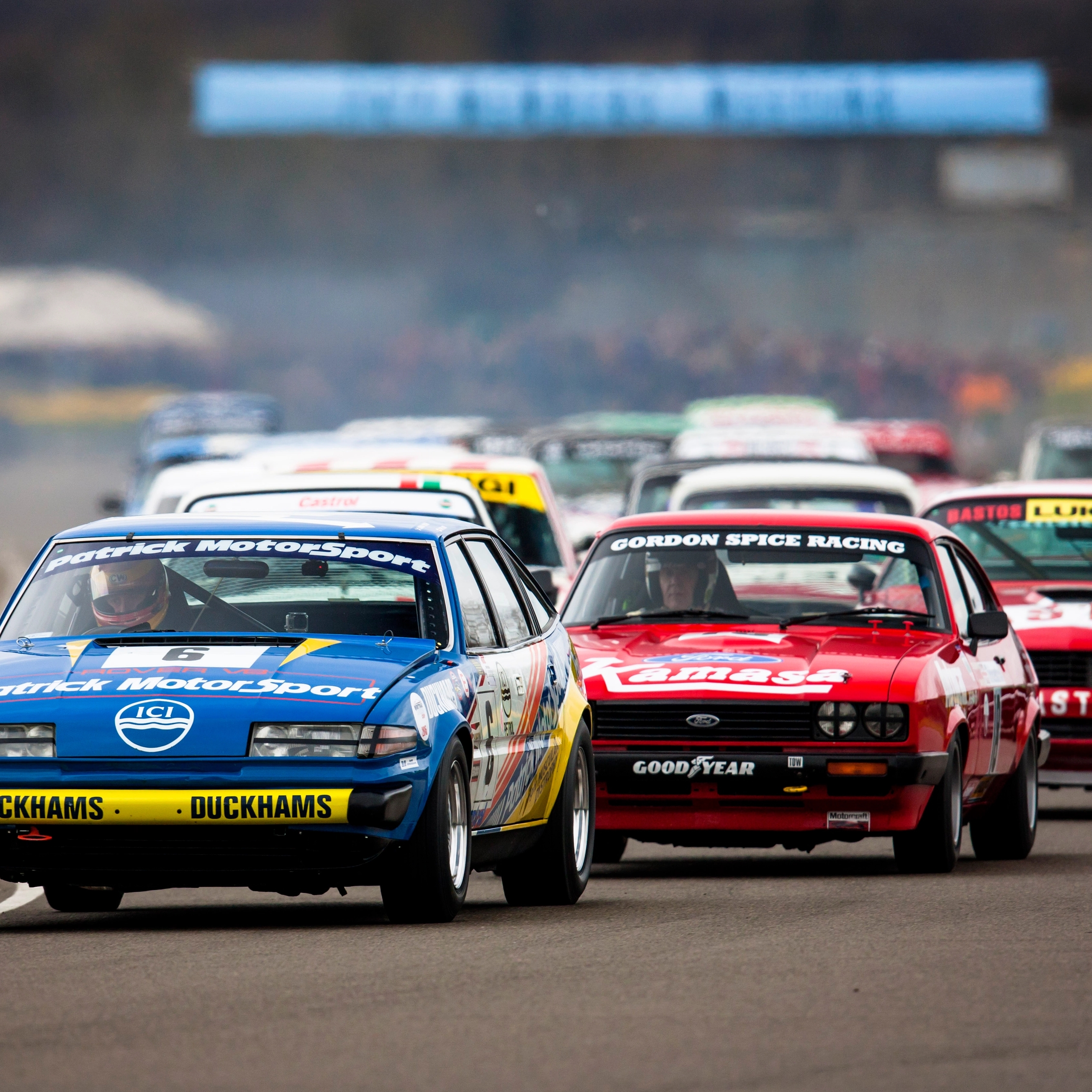 Vintage race cars on a track, with spectators and hills in the background at the 77th Members' Meeting.