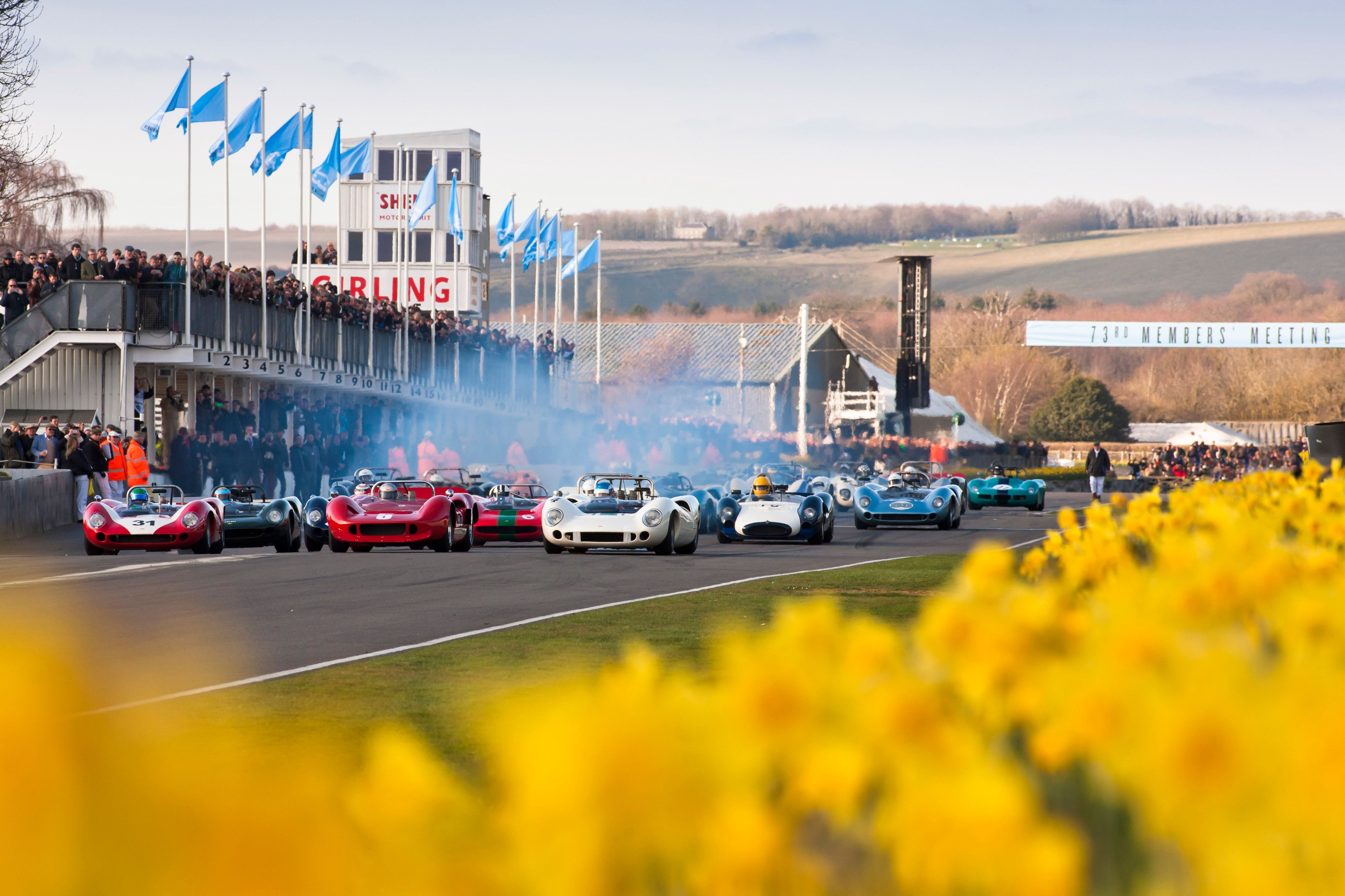 Vintage race cars on a track, with spectators and hills in the background at the 77th Members' Meeting.