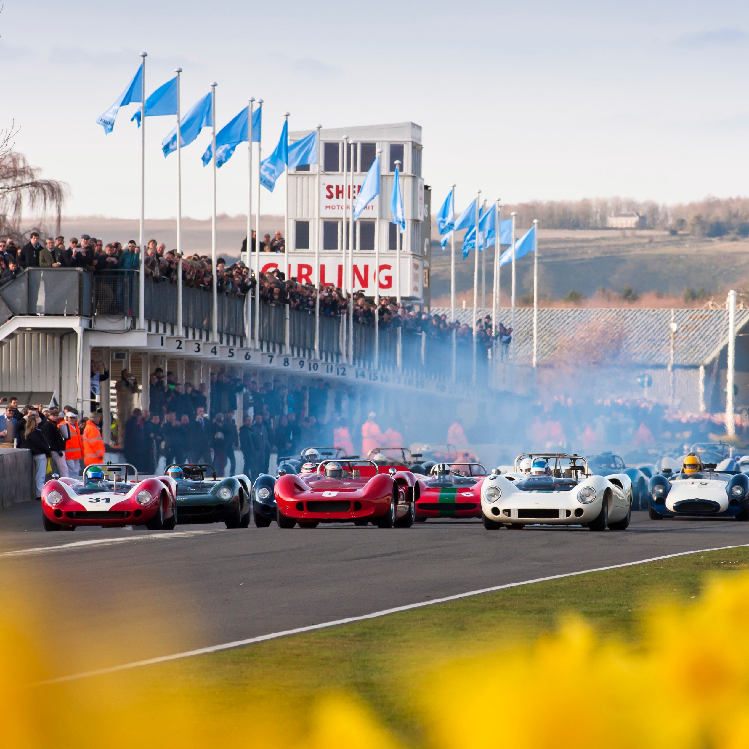Vintage race cars on a track, with spectators and hills in the background at the 77th Members' Meeting.