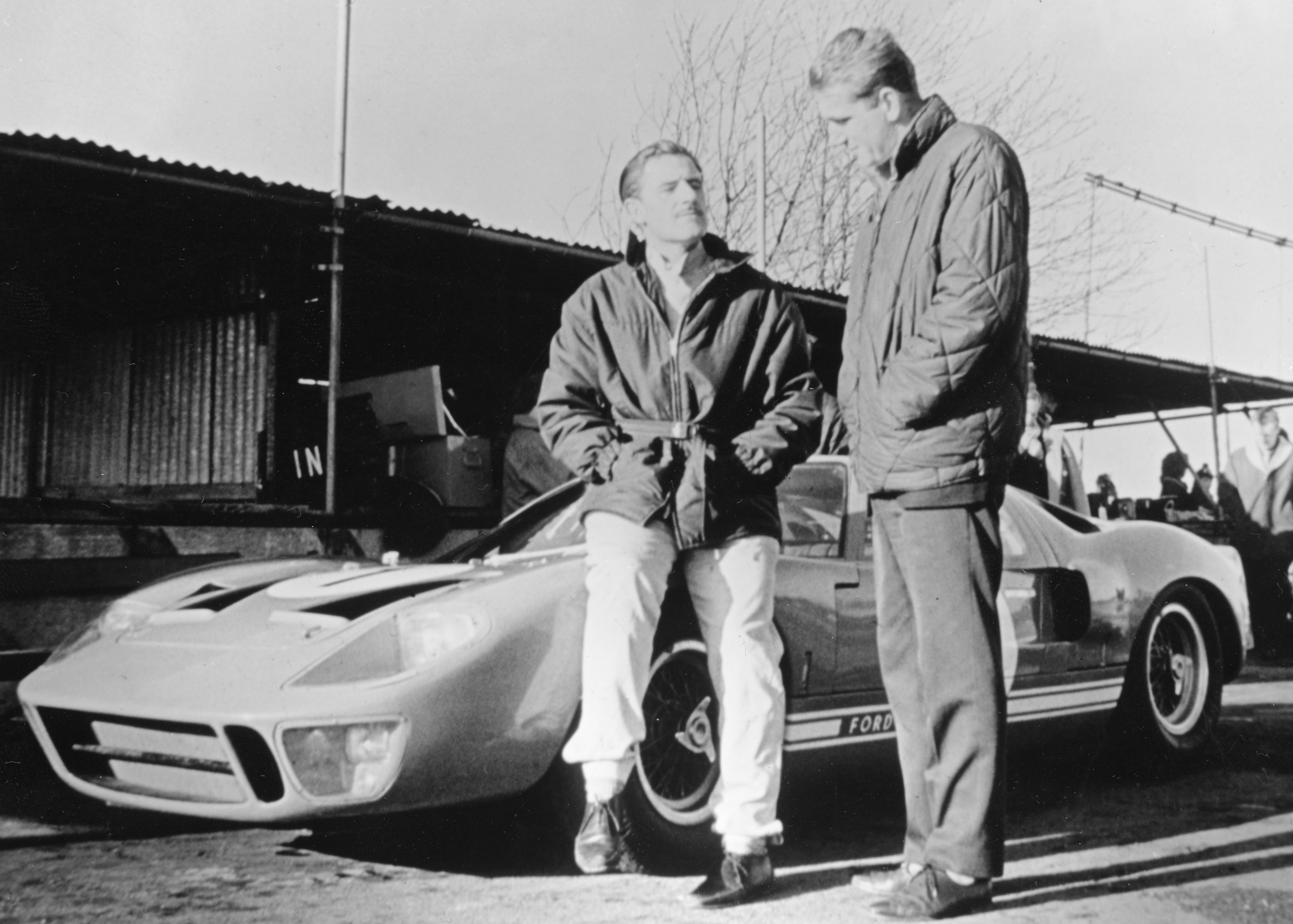 Black and white photo of two men and a Ford GT40 race car; one man sits on the front fender and the other stands next to him.