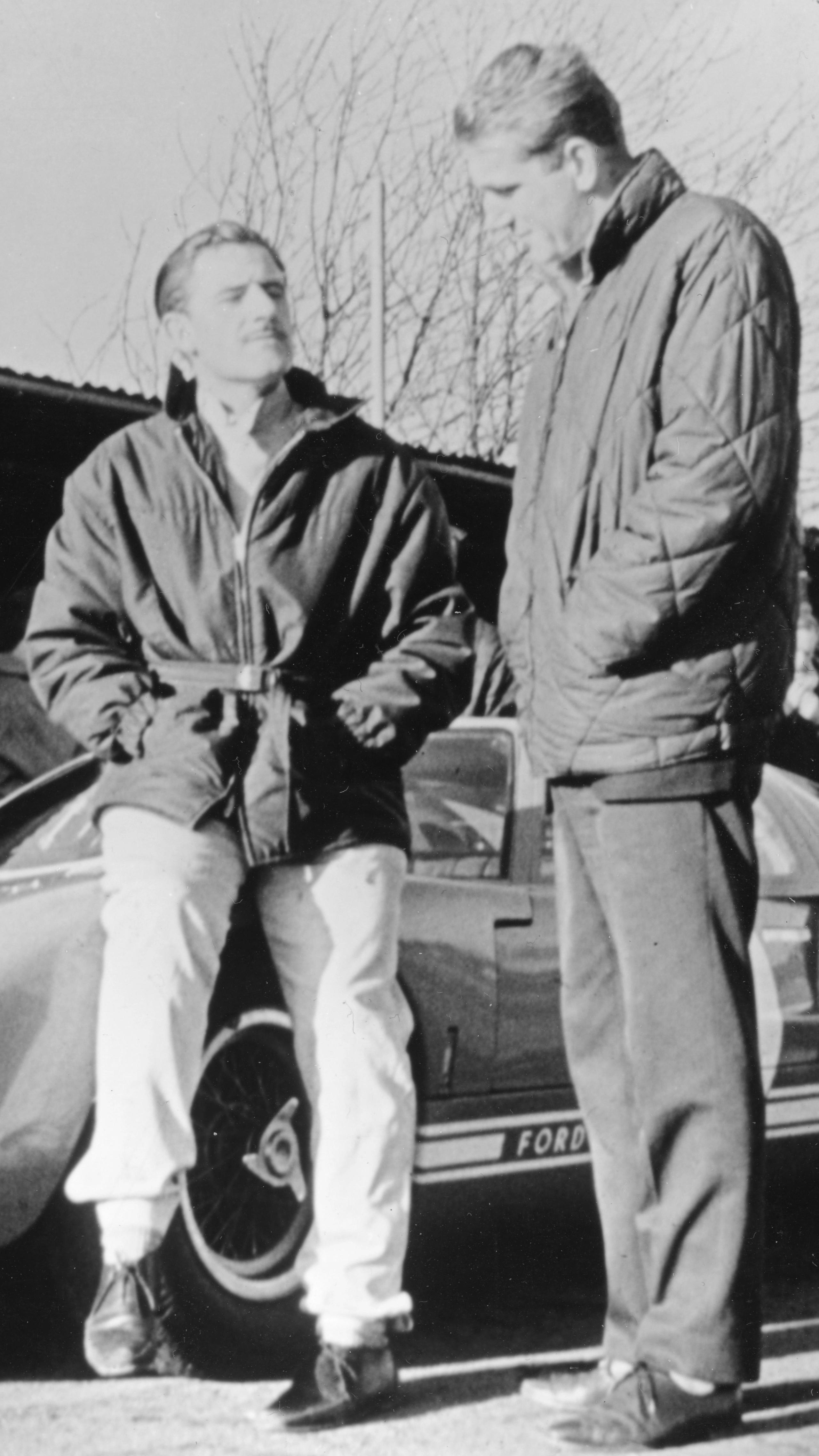 Black and white photo of two men and a Ford GT40 race car; one man sits on the front fender and the other stands next to him.