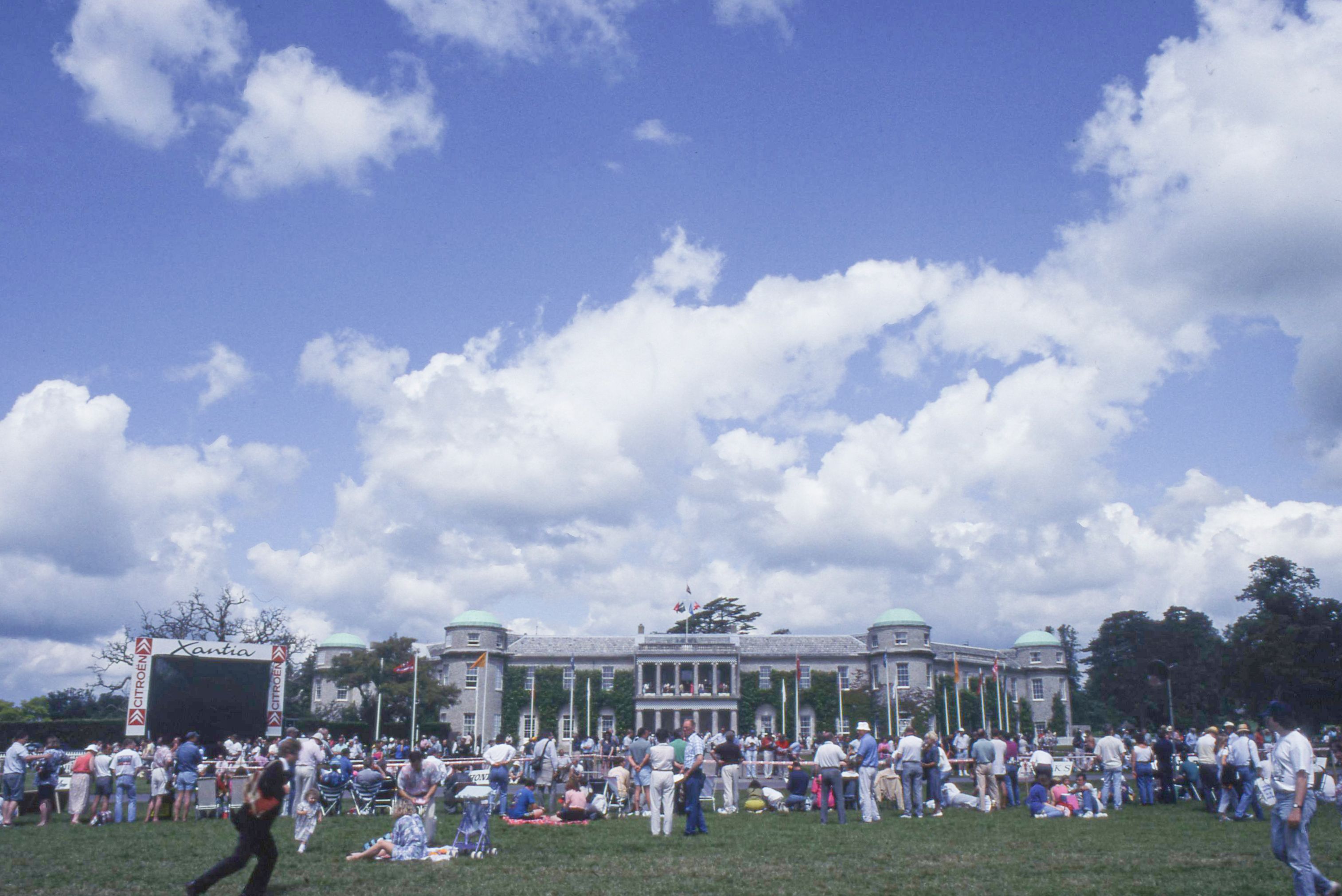 A large crowd gathers on a grassy lawn in front of a grand building with green domes and a big outdoor screen under a cloudy sky.