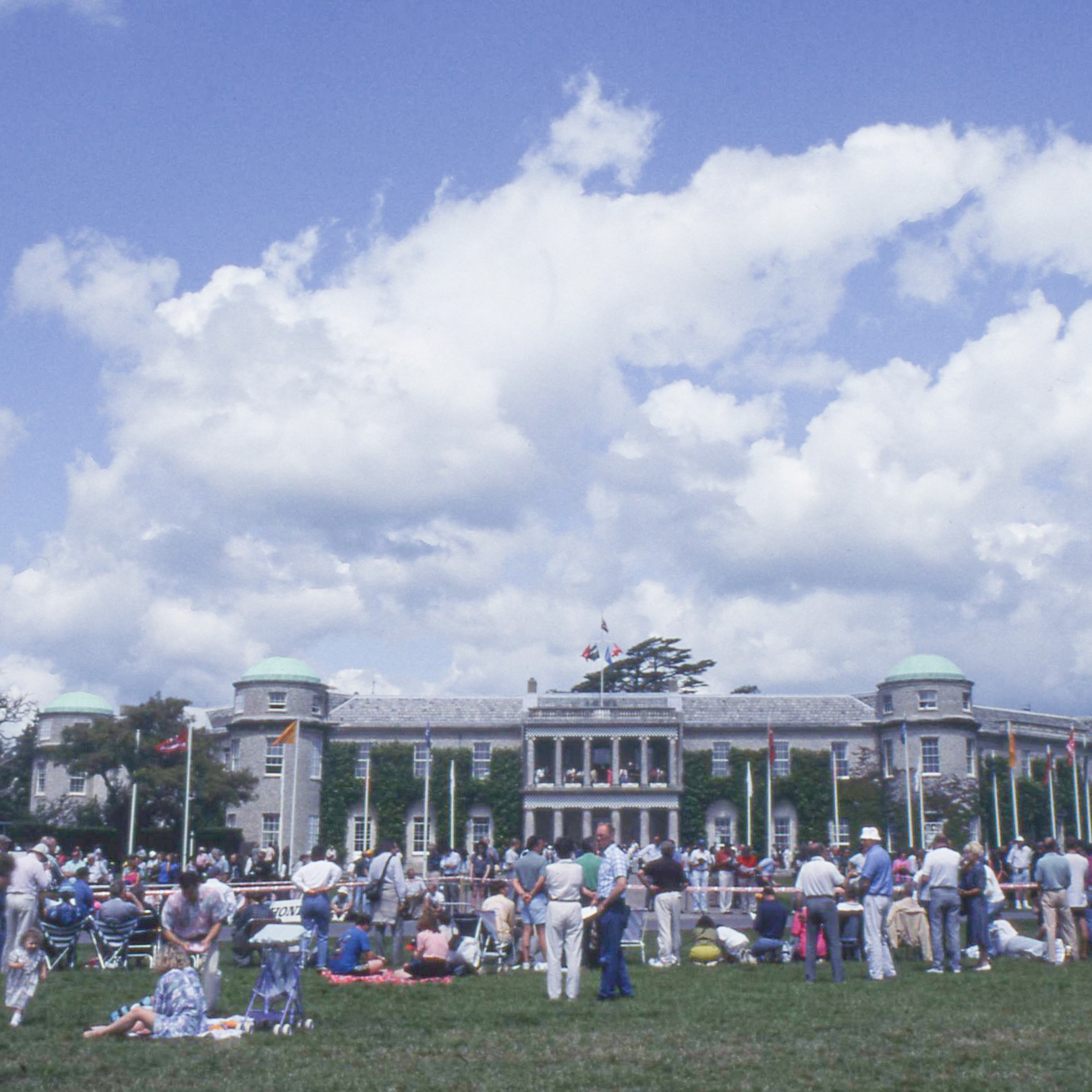 A large crowd gathers on a grassy lawn in front of a grand building with green domes and a big outdoor screen under a cloudy sky.