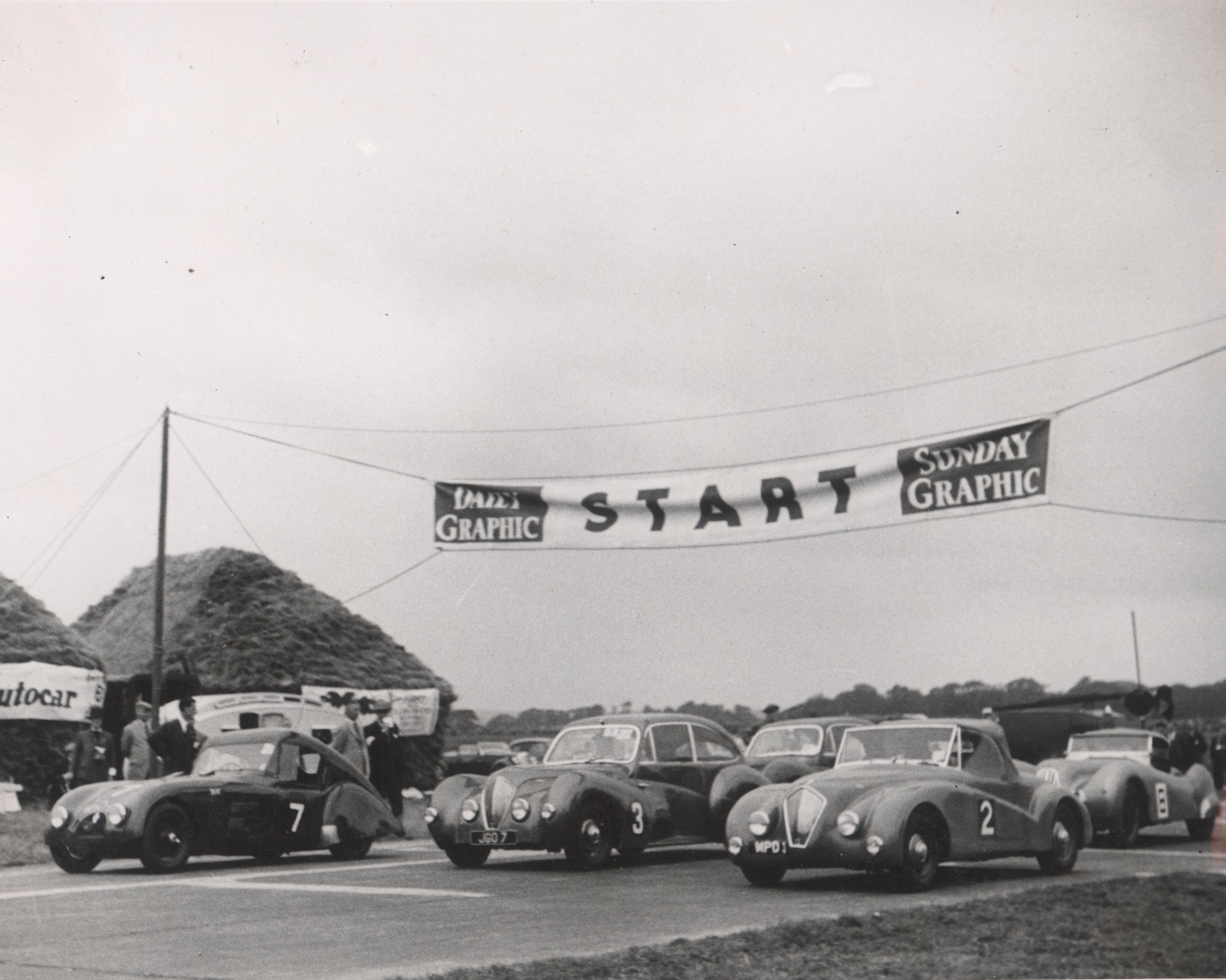 Black and white photograph of vintage sports cars lined up at a race start line under a "START" banner.