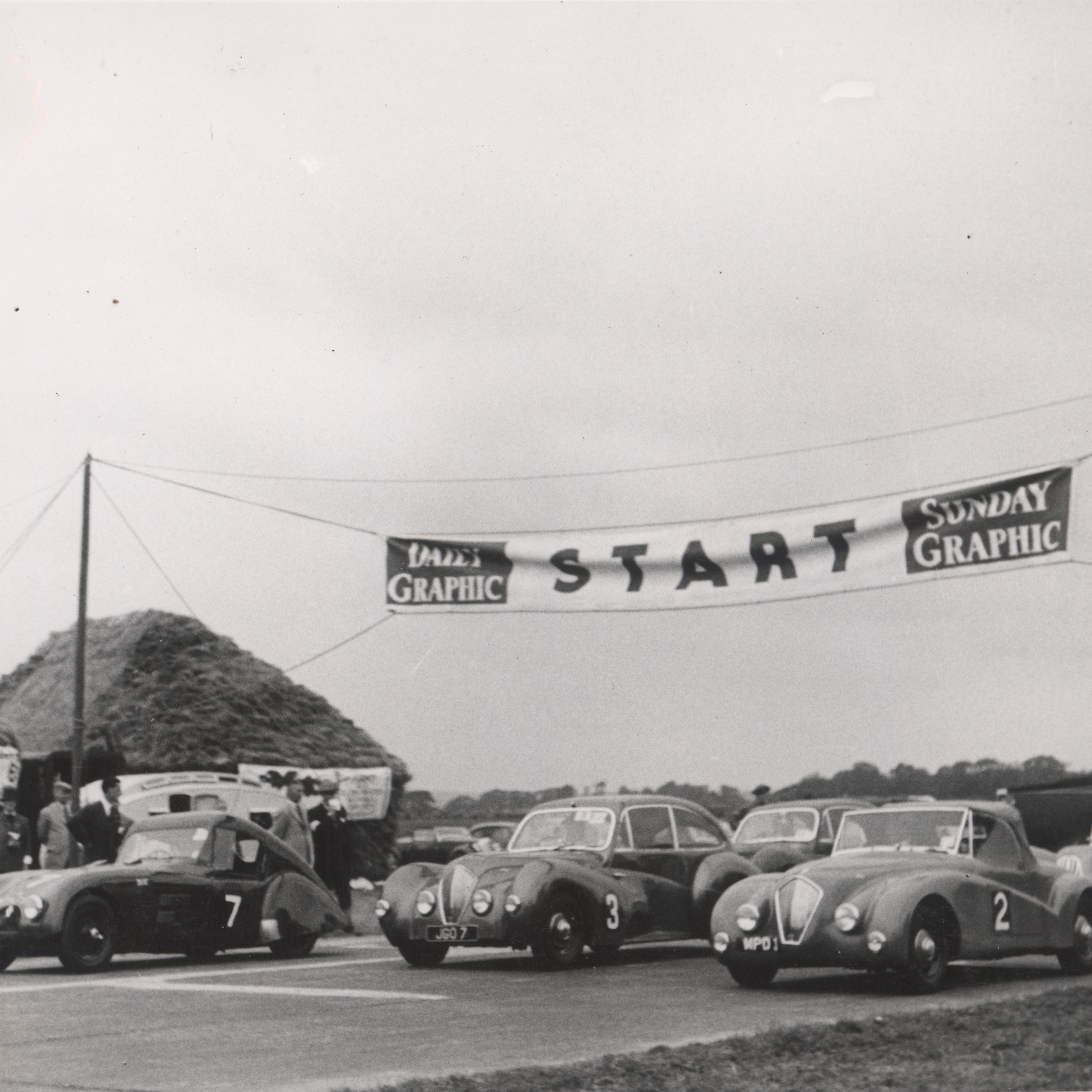 Black and white photograph of vintage sports cars lined up at a race start line under a "START" banner.