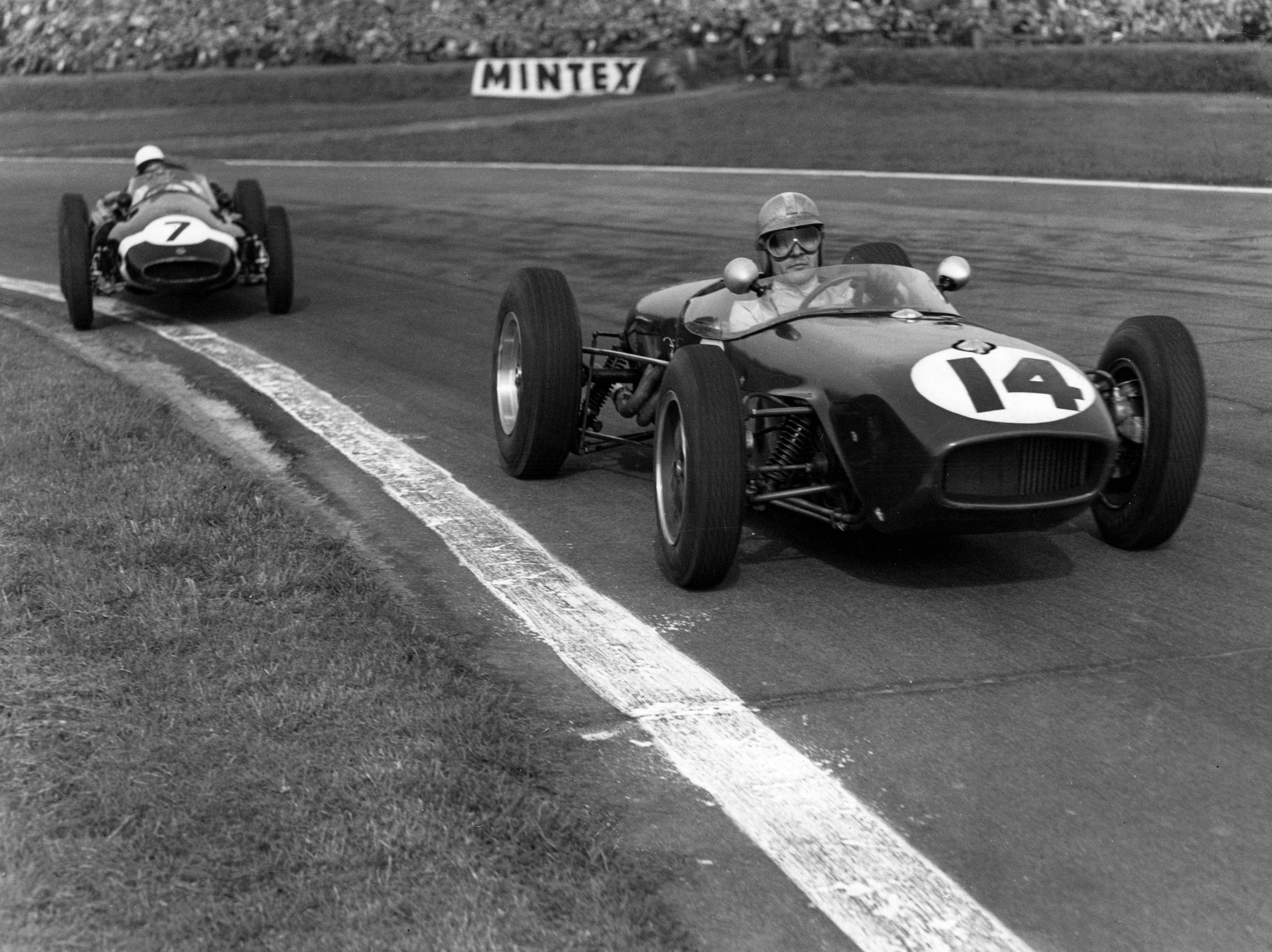 Black and white image of two vintage open-wheel race cars on a track, #14 in the foreground and #7 behind.