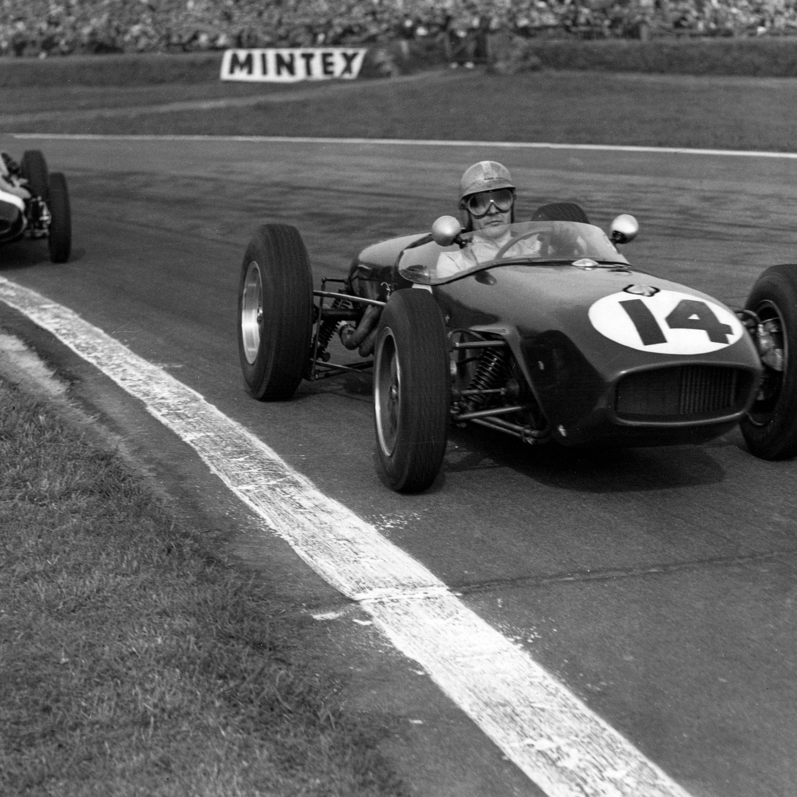 Black and white image of two vintage open-wheel race cars on a track, #14 in the foreground and #7 behind.
