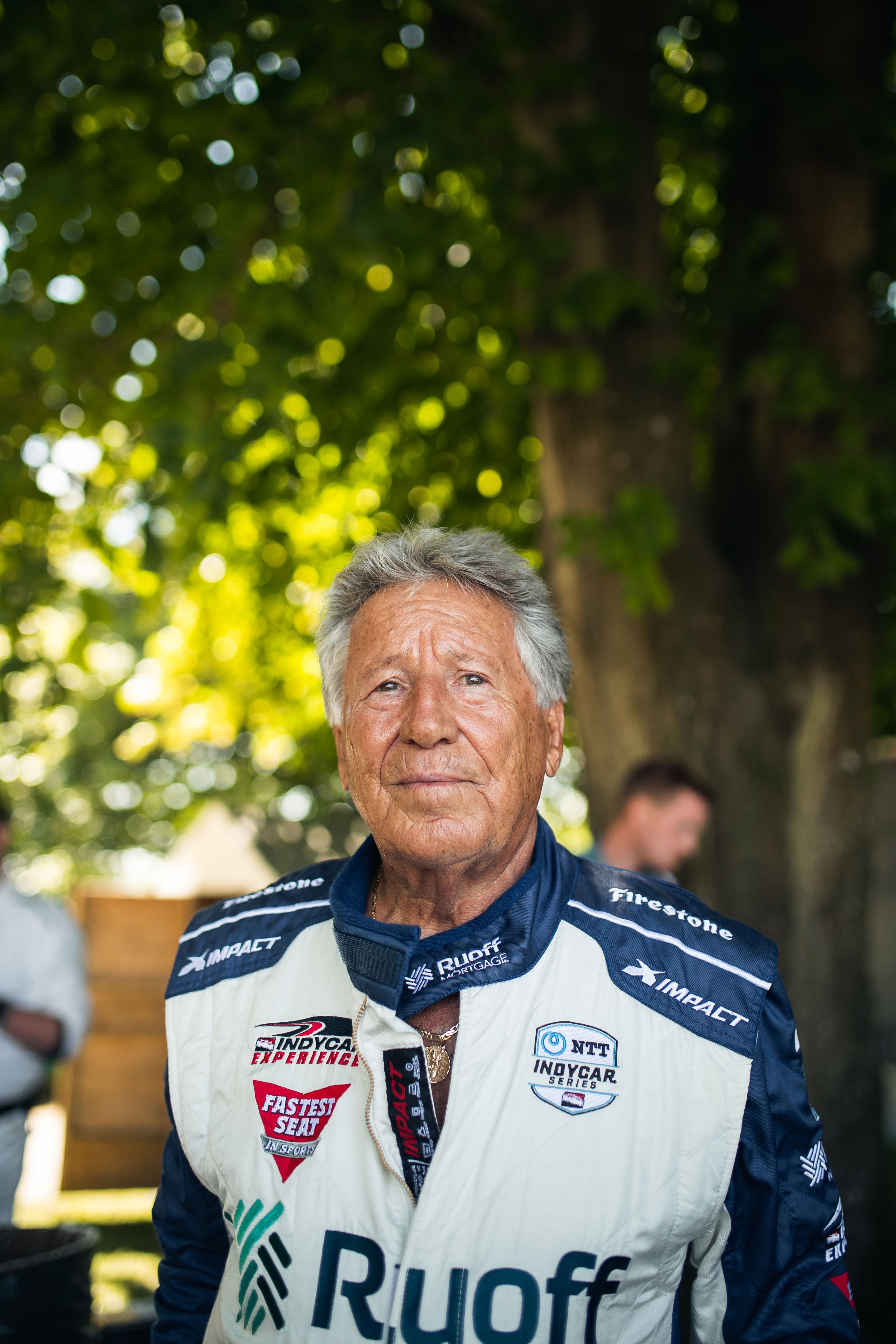 Mario Andretti in a white and blue racing suit smiles at the camera, with blurred green trees in the background.