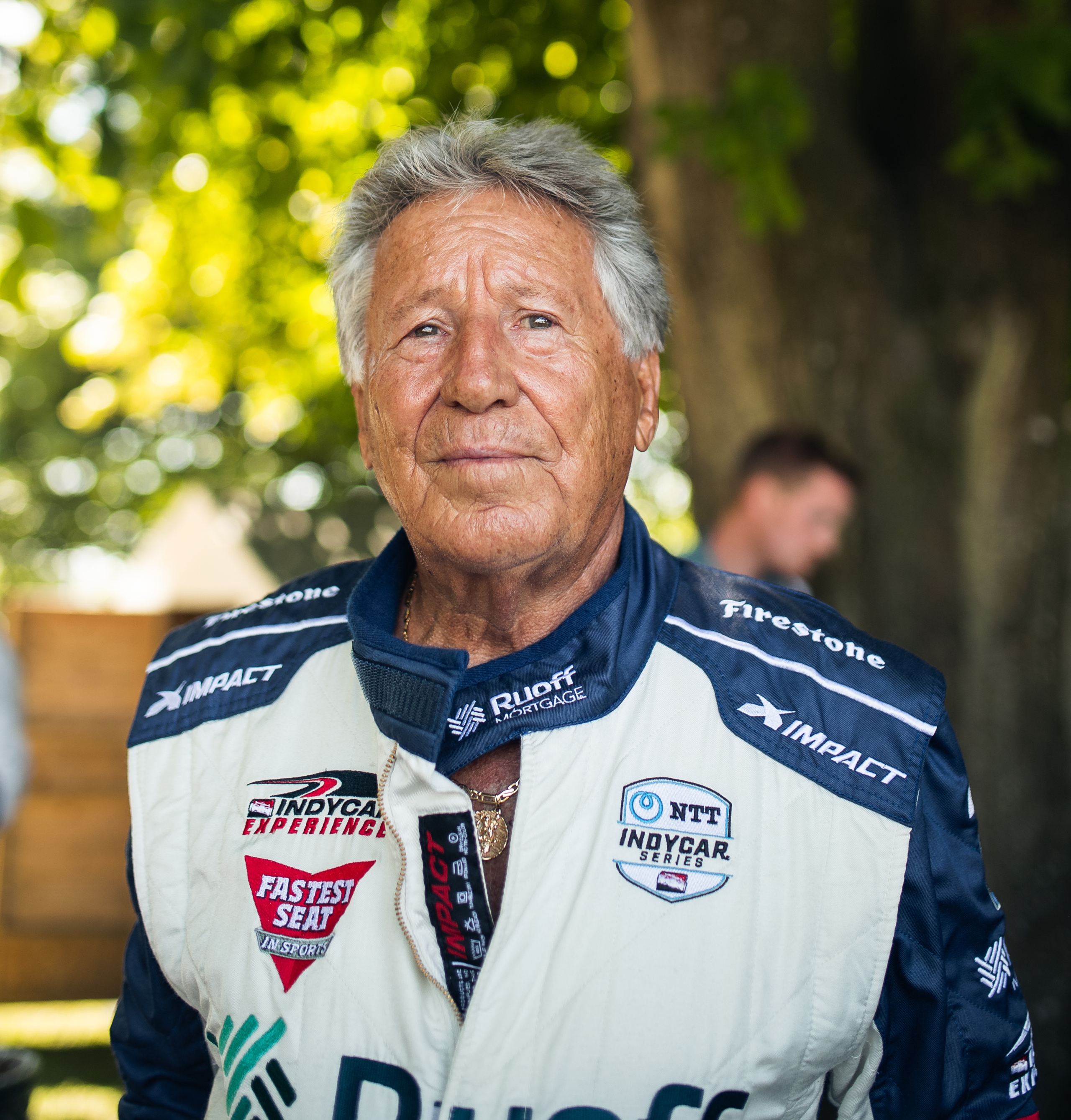 Mario Andretti in a white and blue racing suit smiles at the camera, with blurred green trees in the background.