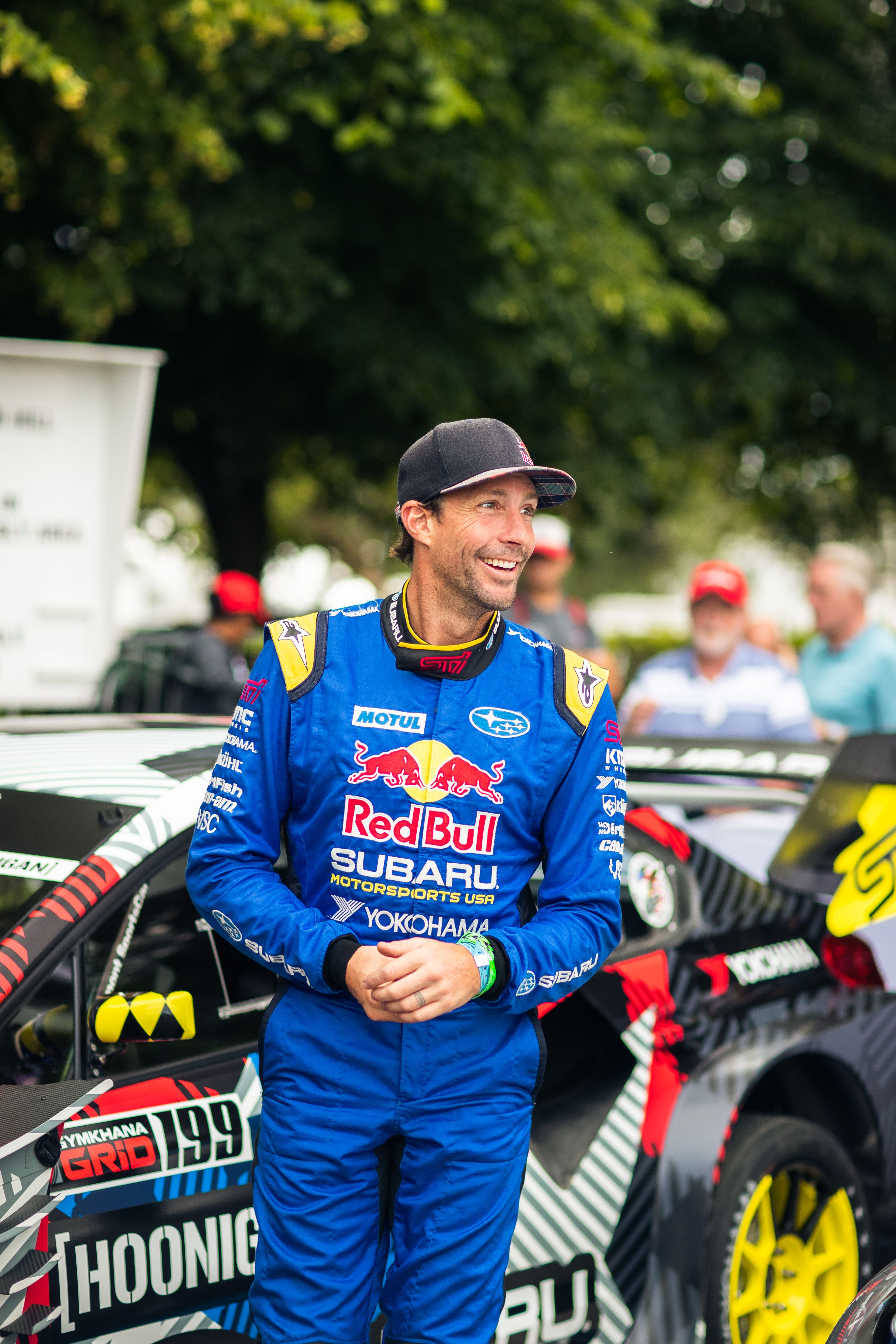 Travis Pastrana in a blue Red Bull Subaru racing suit stands next to a black and white race car.